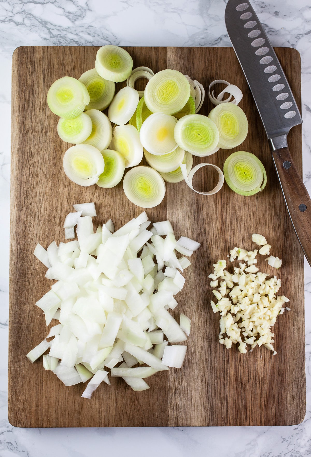 Minced garlic, onions, and sliced leeks on wooden cutting board with knife.