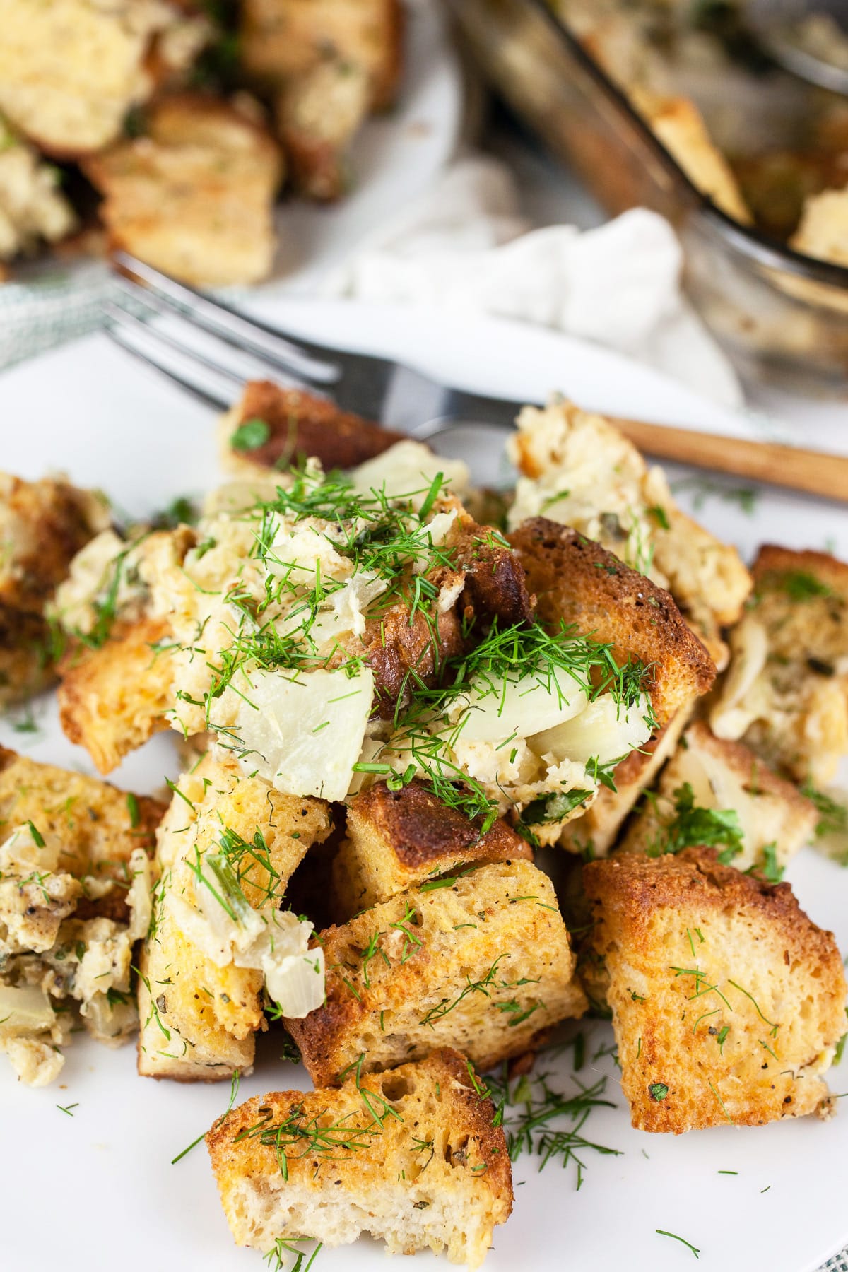 This Savory Fennel and Spinach Stuffing recipe is the perfect side dish for Thanksgiving dinner! It's healthy vegetarian comfort food at its best. This stuffing is made with toasted bread cubes and sauteed fennel. It's baked in the oven and super moist. This homemade stuffing is a unique take on a classic traditional dish. Stuffing from scratch is the best! #stuffing #fennel #spinach #thanksgiving #sides #recipes Fennel herb stuffing on white plate with fork.