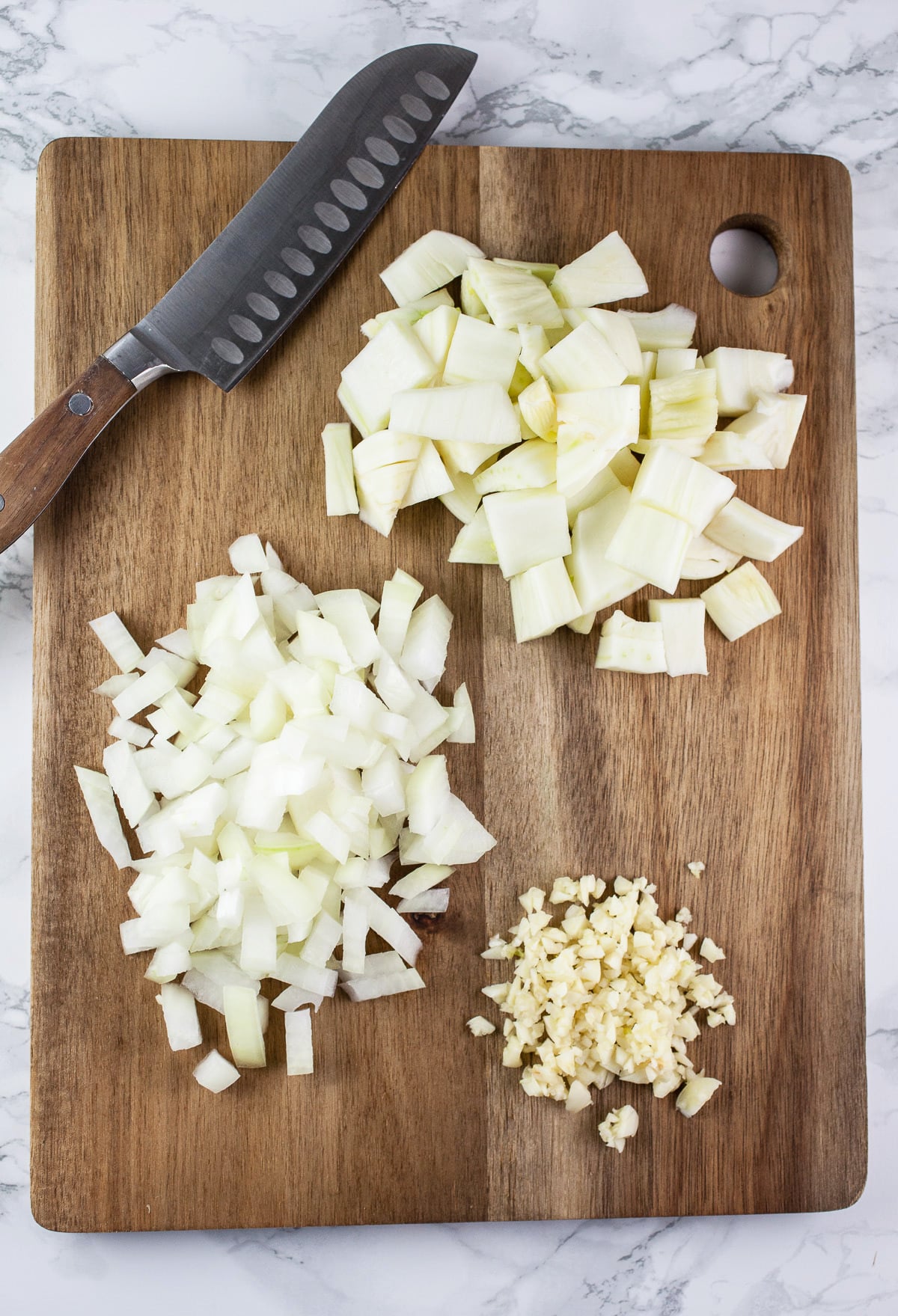 Minced garlic, onions, and fennel on wooden cutting board with knife.