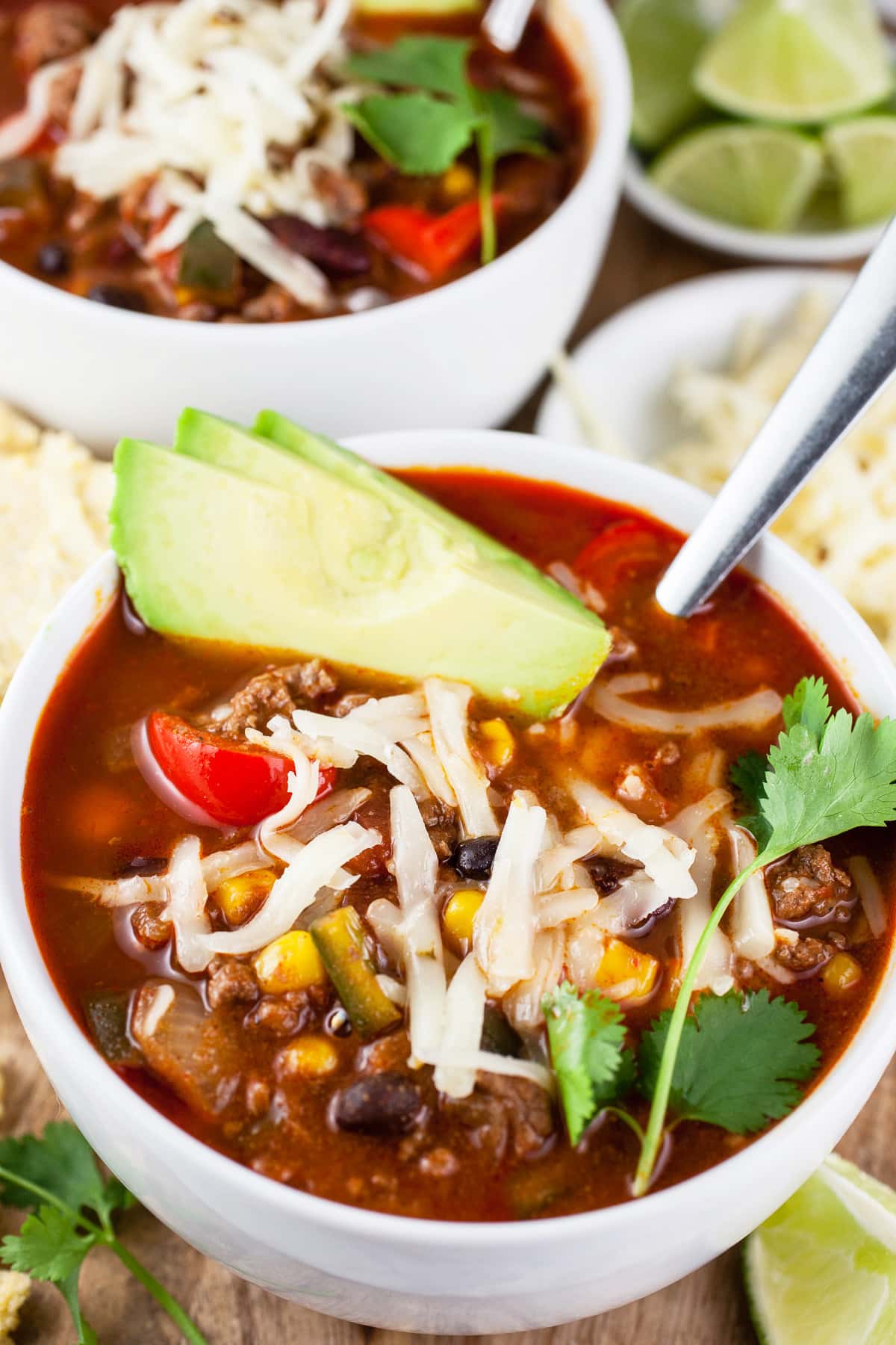 Ground beef chili with cheese, avocado, cilantro, and limes in bowls.