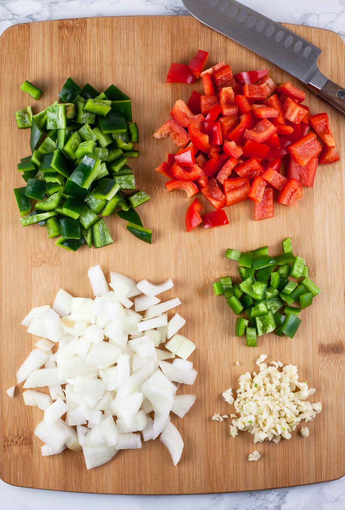 Minced garlic, onions, jalapeno, red bell pepper, and poblano pepper on wooden cutting board with knife.
