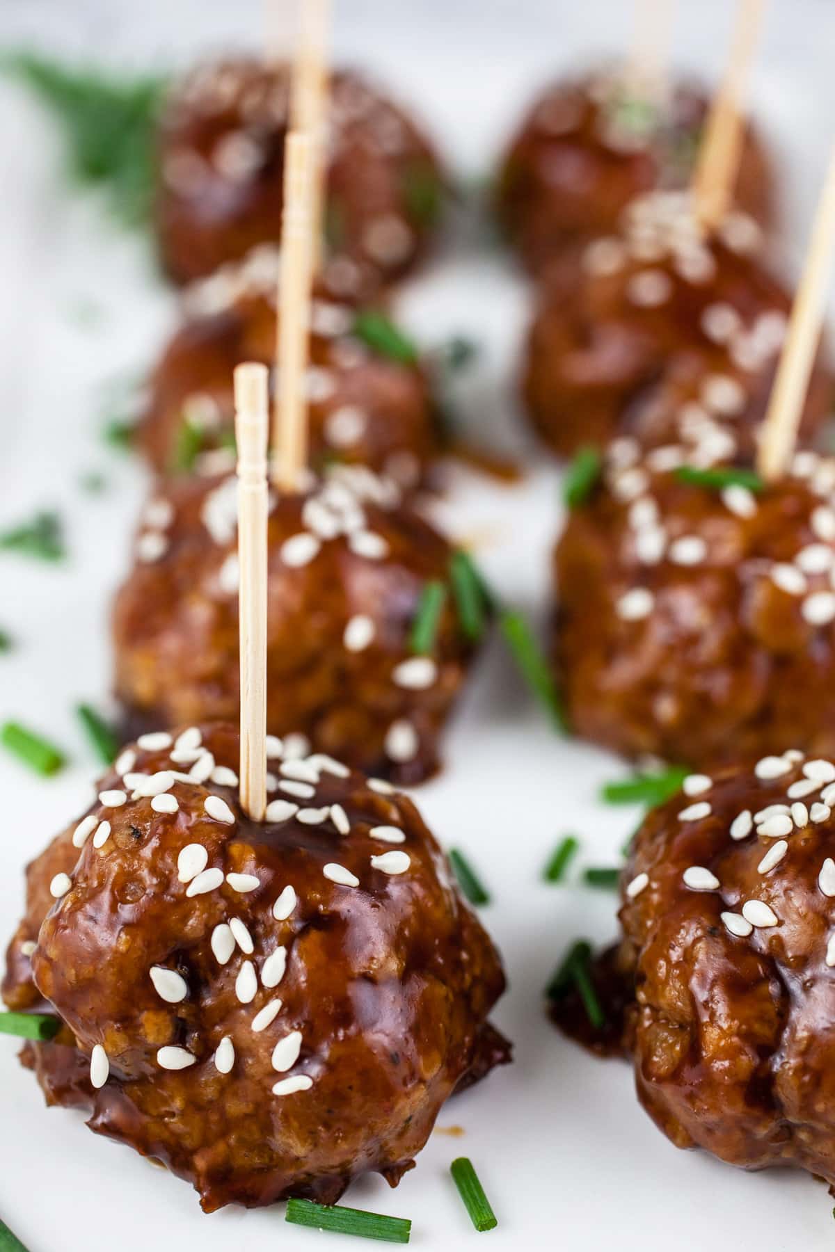Teriyaki meatballs with sesame seeds and chives on white platter with toothpicks.