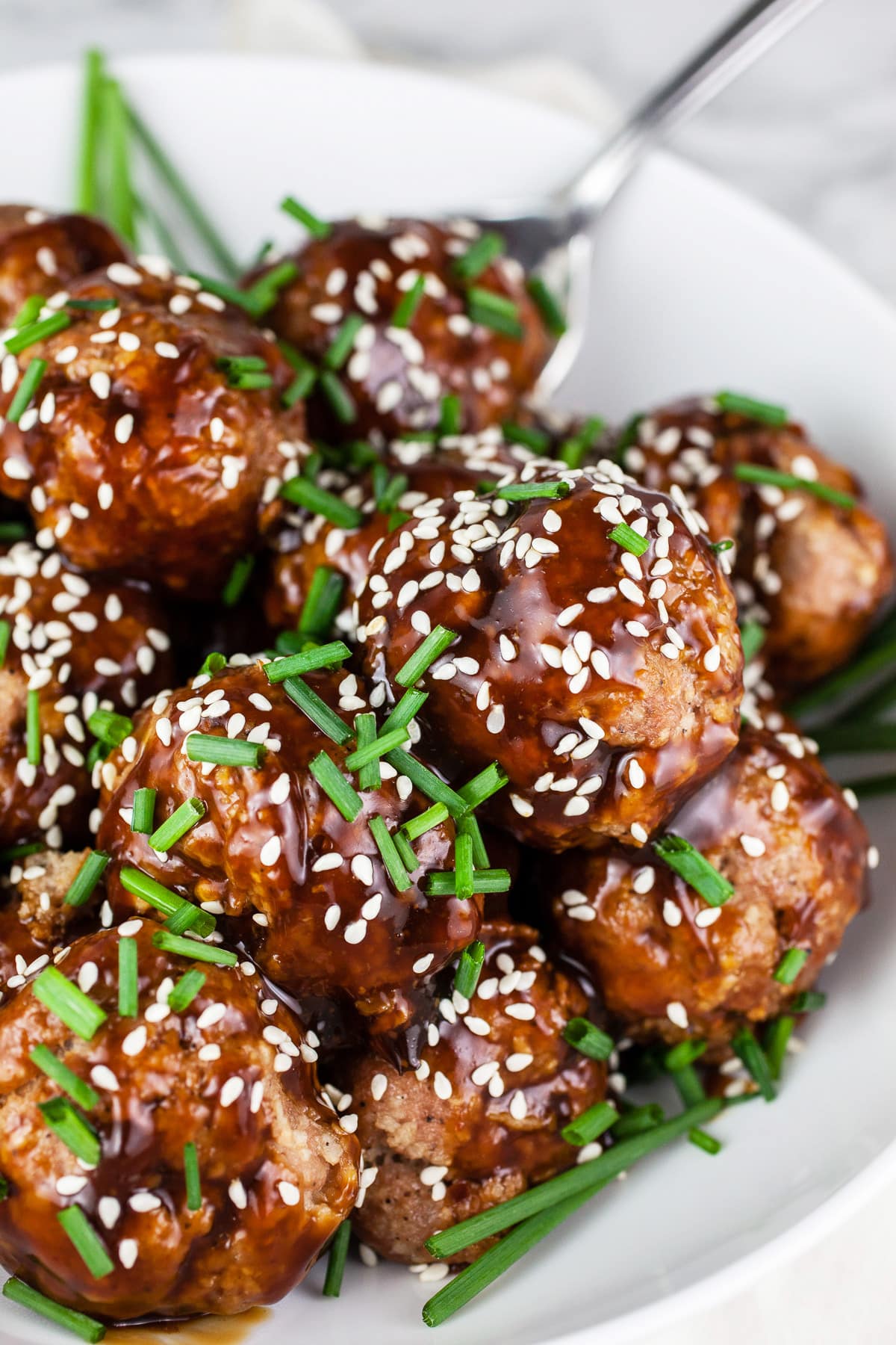 These Crockpot Teriyaki Meatballs are easy to make and full of flavor! These Asian meatballs are made with turkey, baked in the oven, and covered with a sticky sweet and savory sauce. They're warmed in the slow cooker so they're great for holiday parties like Christmas and New Year's Eve. Also great for Game Day. They're gluten free as well! #crockpot #meatballs #turkey #teriyaki #glutenfree Teriyaki turkey meatballs with sesame seeds and chives in white bowl.