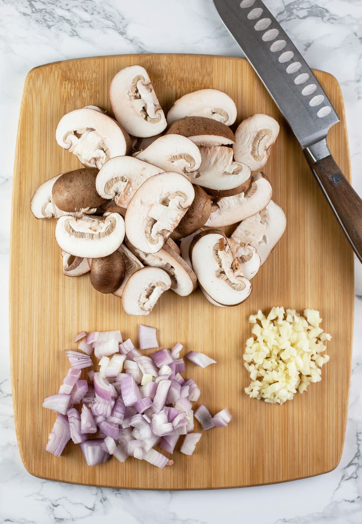 Minced garlic, shallots, and sliced mushrooms on wooden cutting board with knife.
