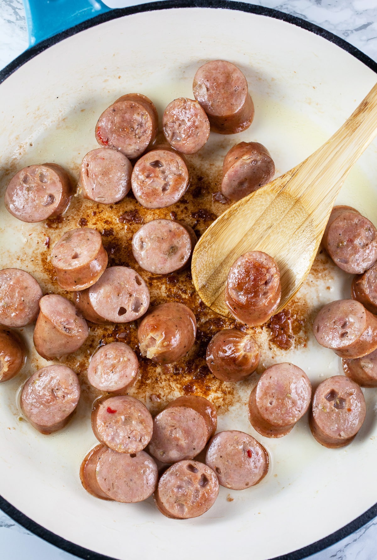 Sliced chicken sausages sautéed in skillet with wooden spoon.