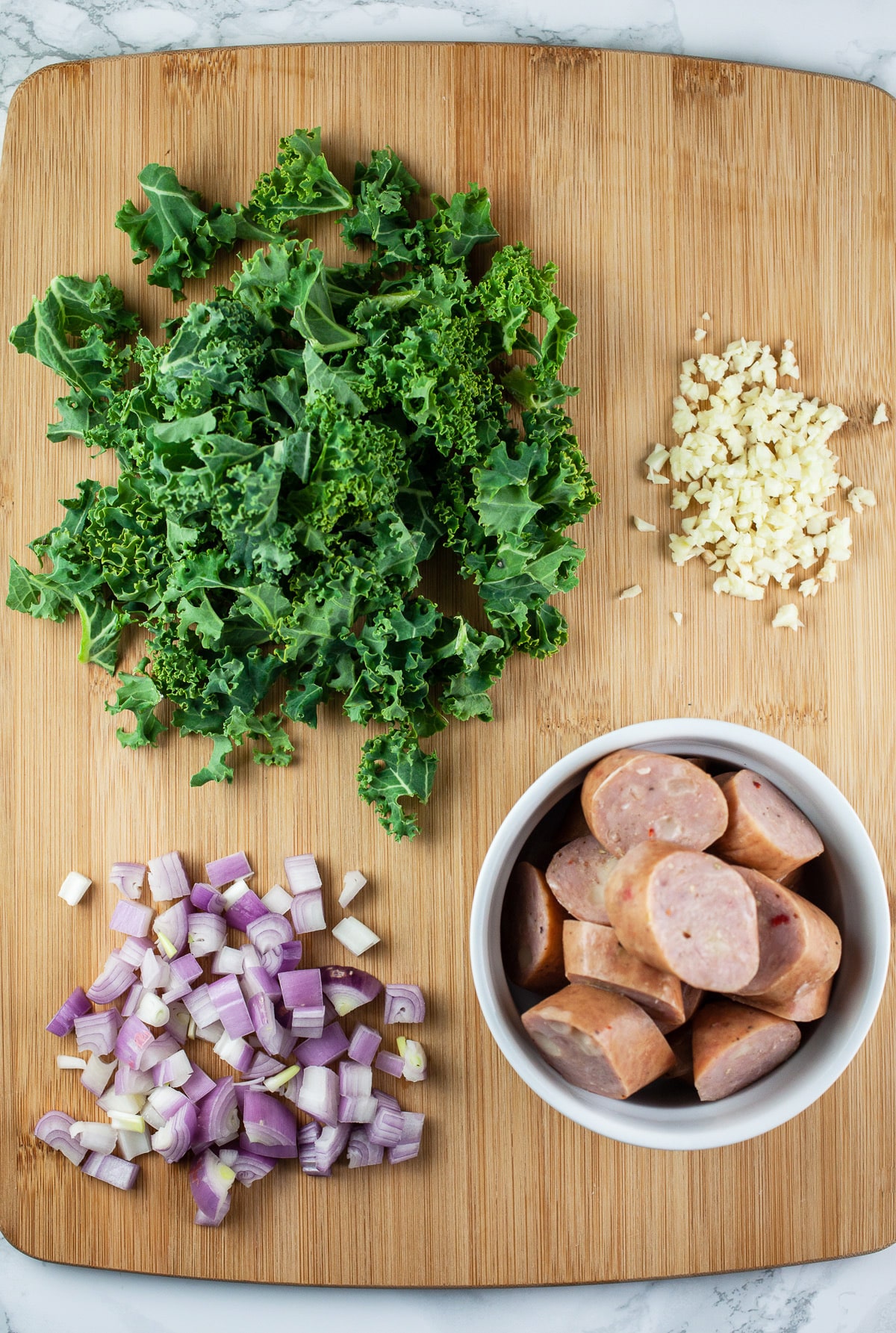 Minced garlic, shallots, and kale and sliced chicken sausages on wooden cutting board.