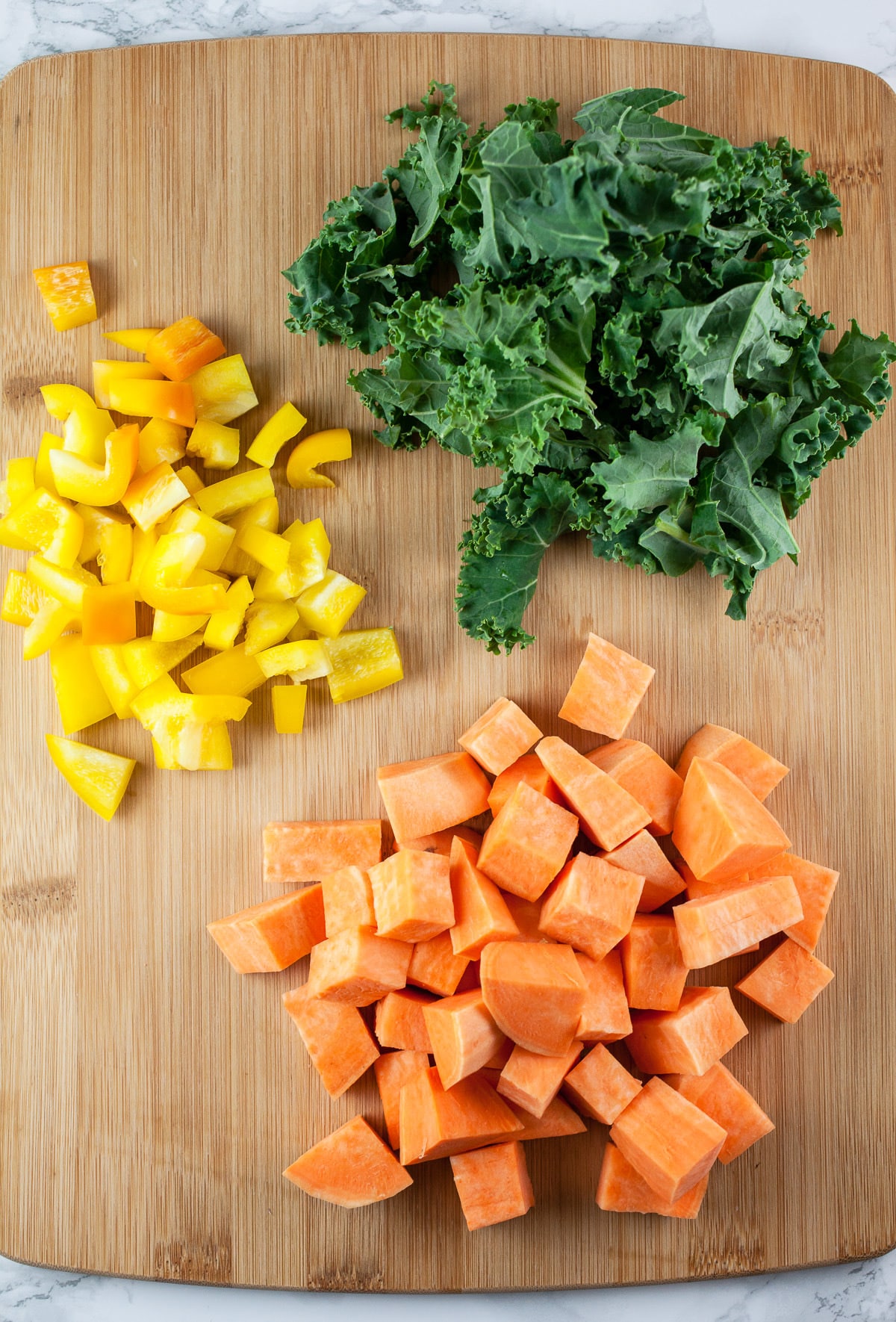 Diced sweet potatoes, yellow bell peppers, and chopped kale on wooden cutting board.