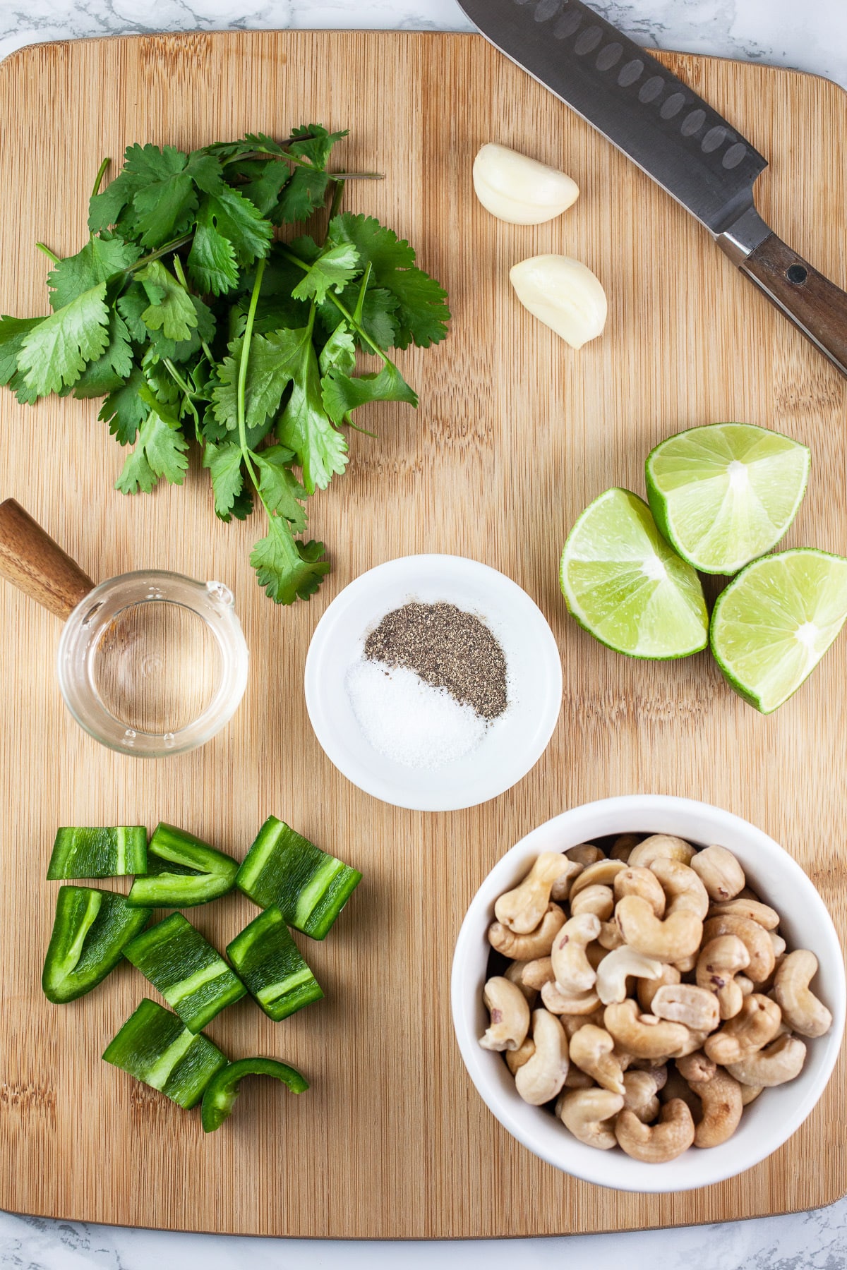 Cashews, jalapeno pepper, water, cilantro, garlic, limes, salt, and pepper on wooden cutting board.