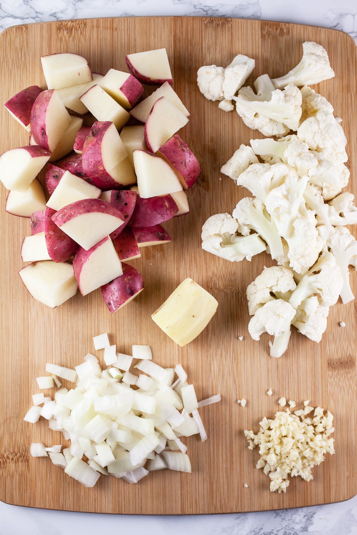 Minced garlic, onions, diced red potatoes, cauliflower florets, and ginger root on wooden cutting board.