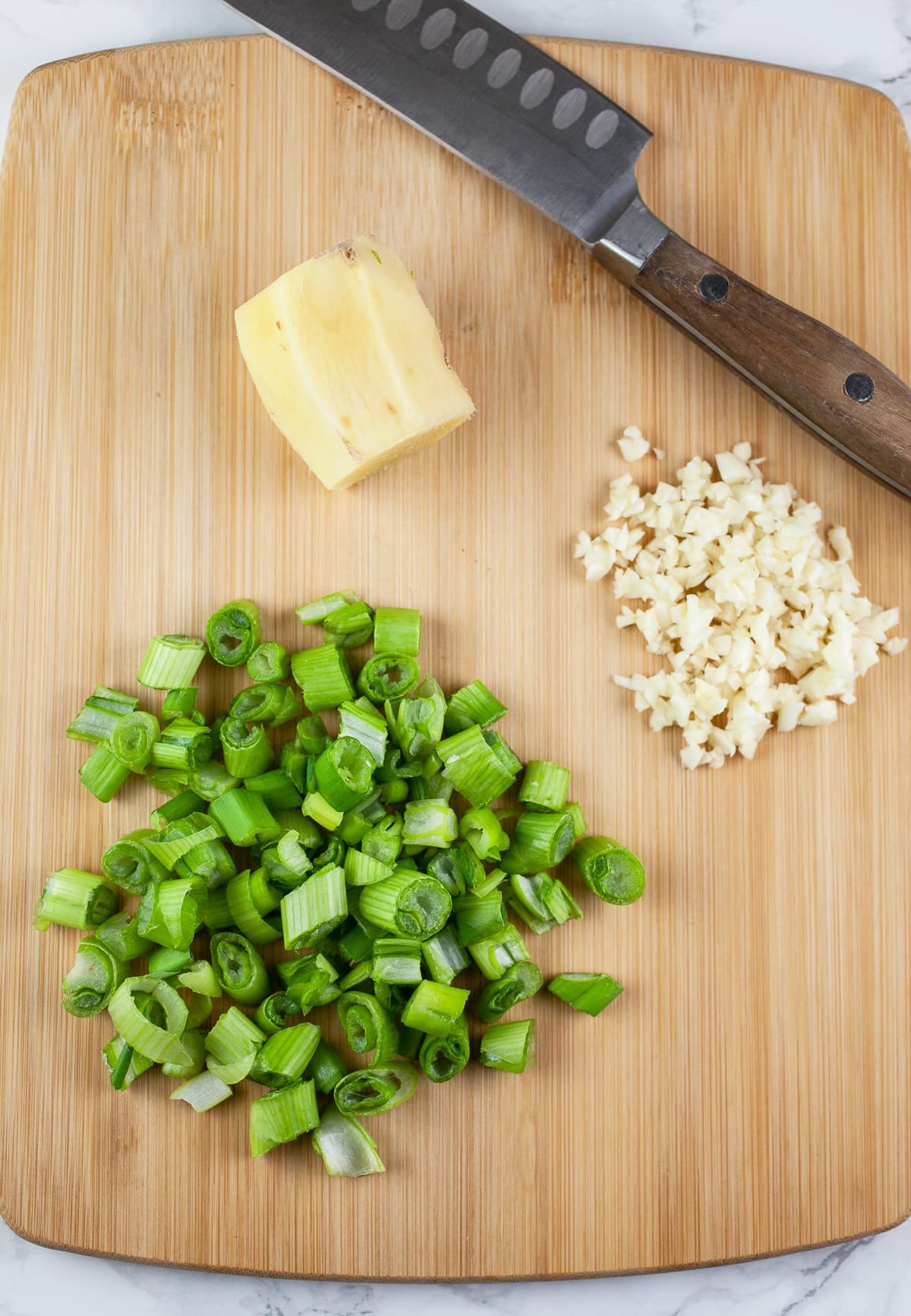 Minced scallions and garlic and ginger root on wooden cutting board with knife.