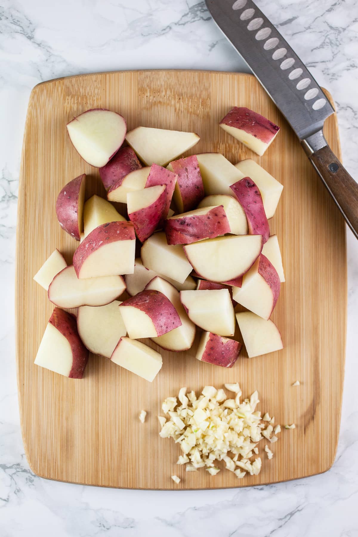 Minced garlic and diced red potatoes on wooden cutting board with knife.