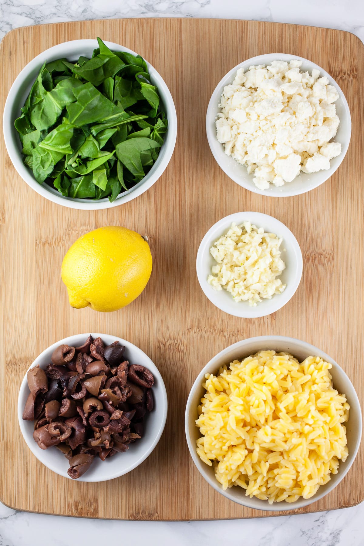 Cooked orzo, Kalamata olives, spinach, feta cheese, garlic, and lemon on wooden cutting board.