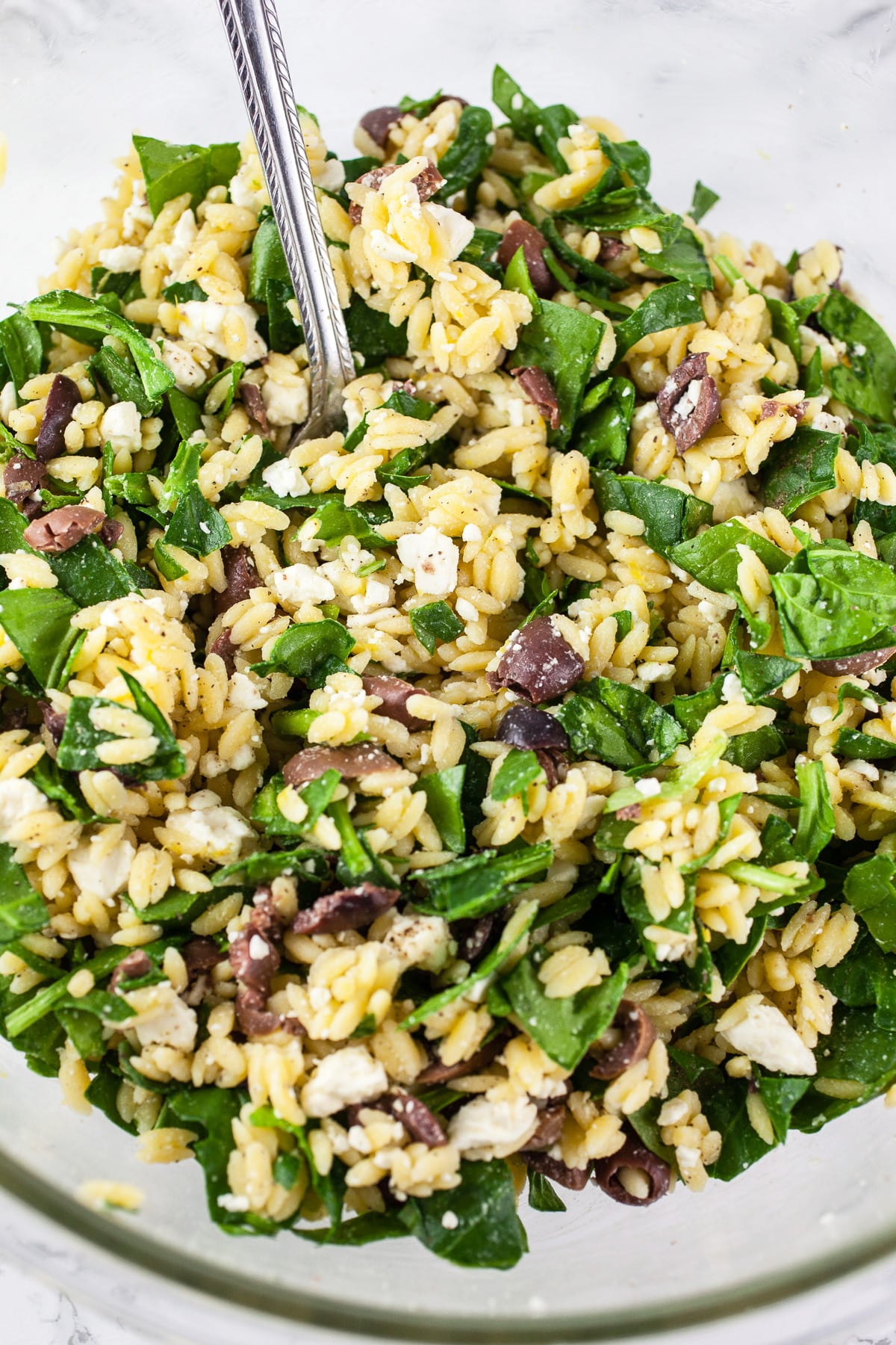 Cooked orzo, spinach, Kalamata olive, feta cheese, and lemon zest combined in large glass bowl with spoon.