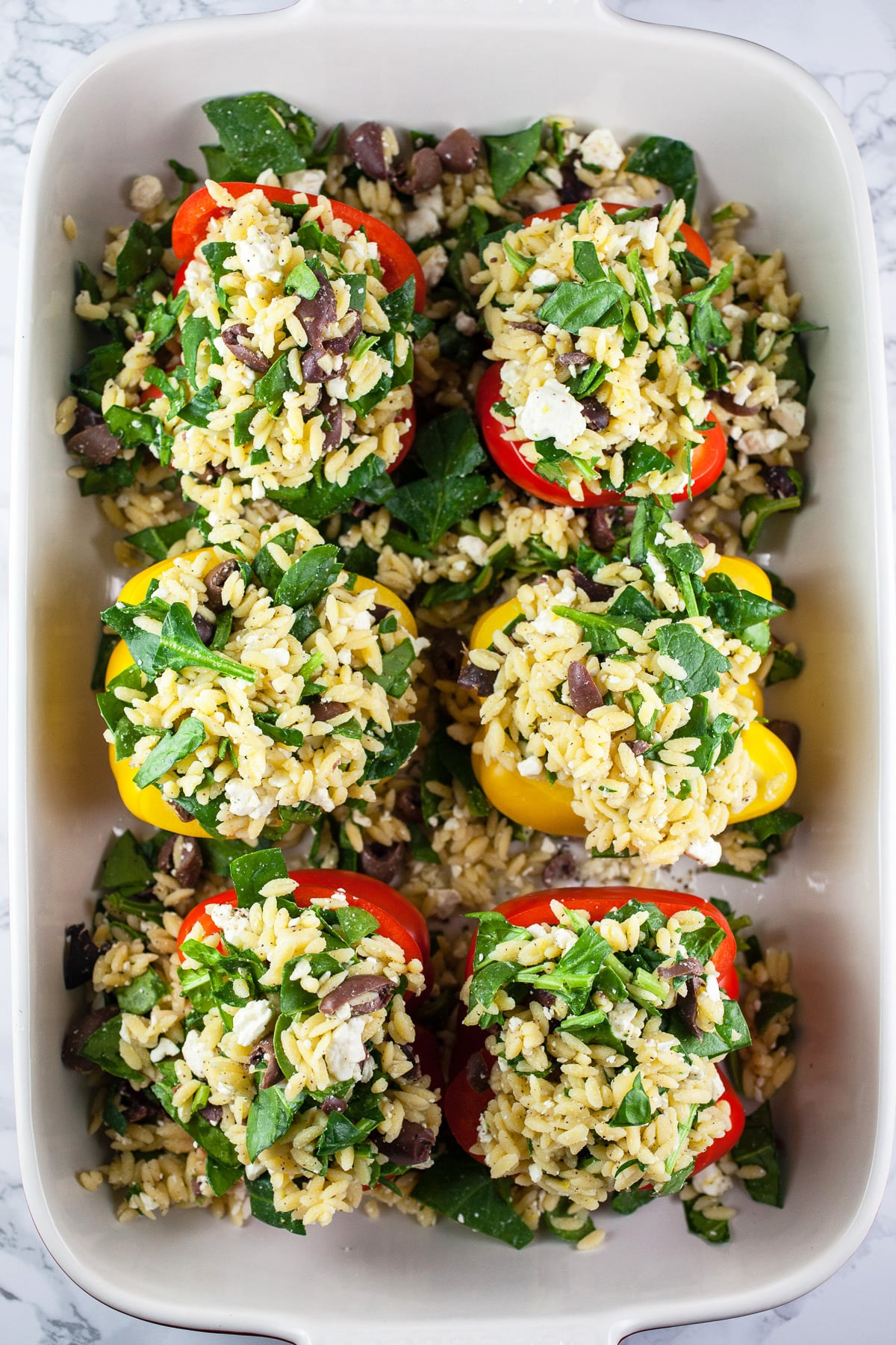 Uncooked bell peppers stuffed with orzo filling in ceramic baking dish.