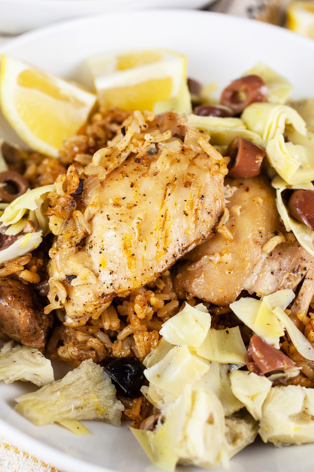 Mediterranean chicken thighs and rice with artichokes, Kalamata olives, and lemon wedges in white bowl.