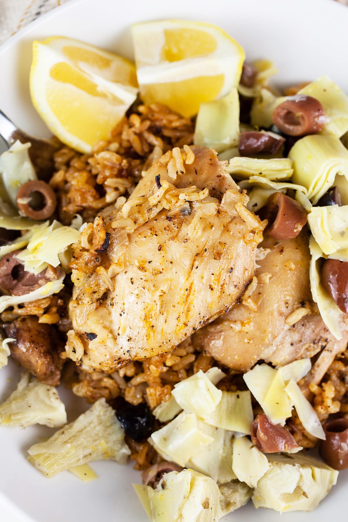 Chicken thighs and rice with artichokes, olives, and lemon wedges in white bowl.