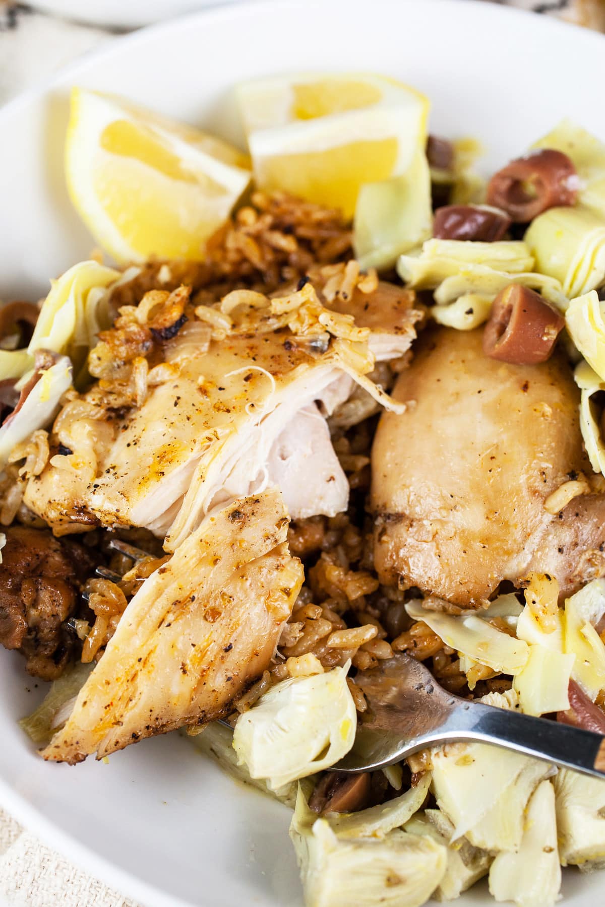 Chicken thighs and rice with artichokes, olives, and lemon wedges in white bowl with fork.