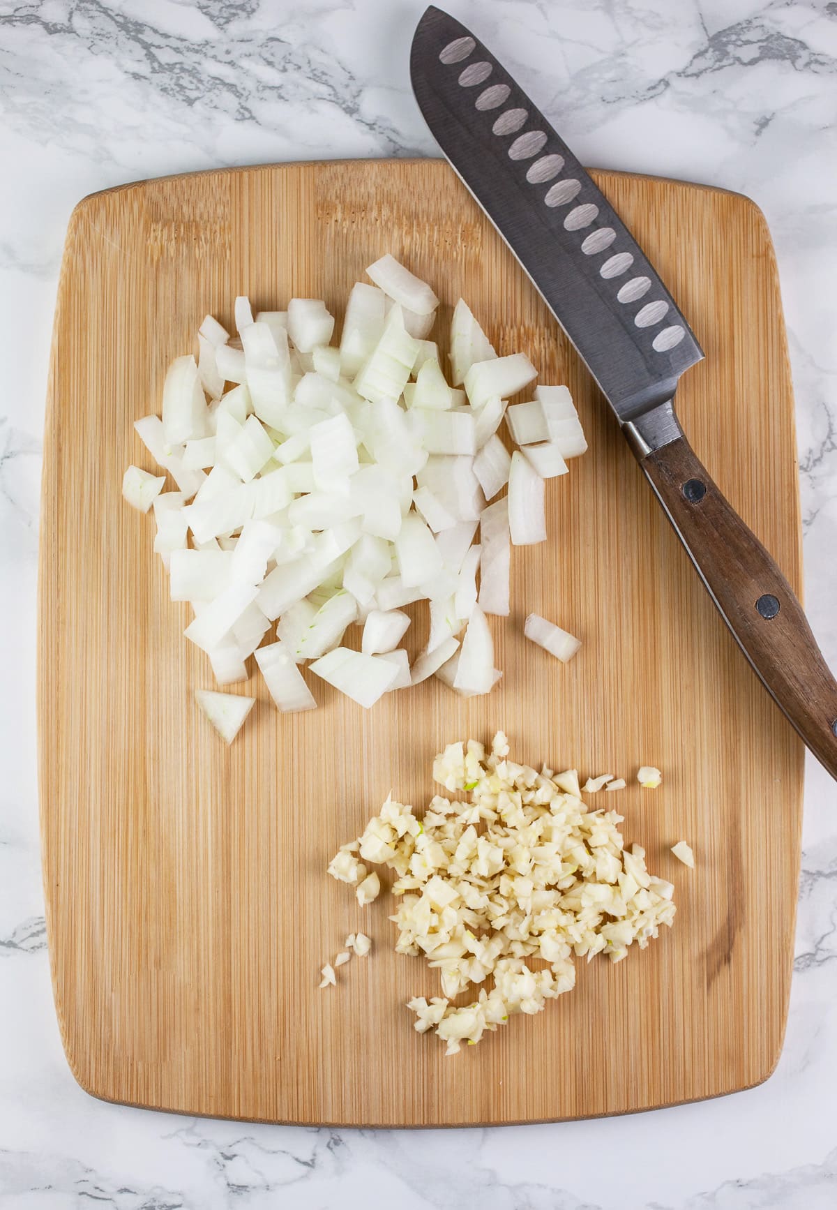 Minced garlic and onions on wooden cutting board with knife.