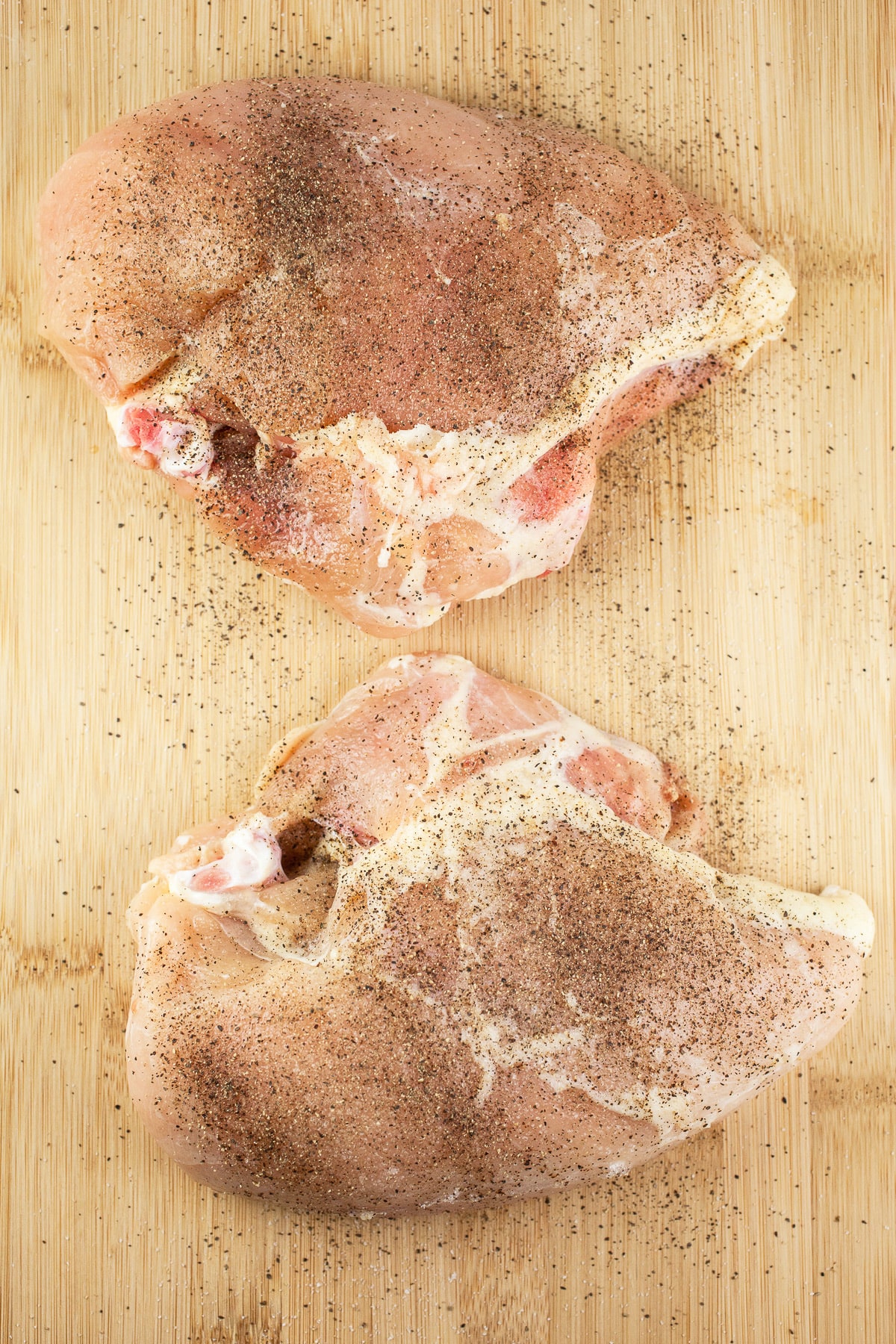 Raw split chicken breasts with salt and pepper on wooden cutting board.