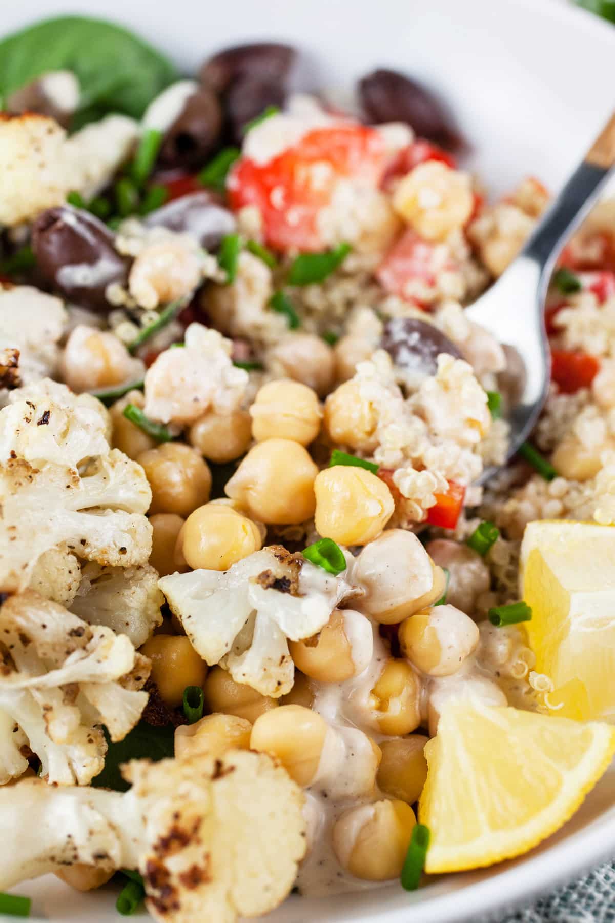 Mediterranean chickpea quinoa bowl with cauliflower, red bell peppers, spinach, Kalamata olives, tahini dressing, and lemon wedges.