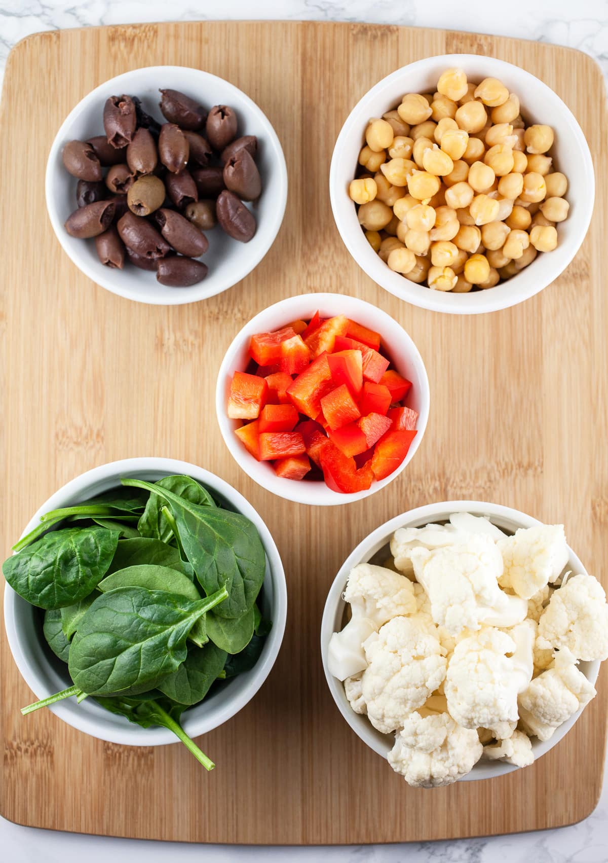 Cauliflower florets, spinach, Kalamata olives, chickpeas, and diced red bell peppers in bowls on wooden cutting board.