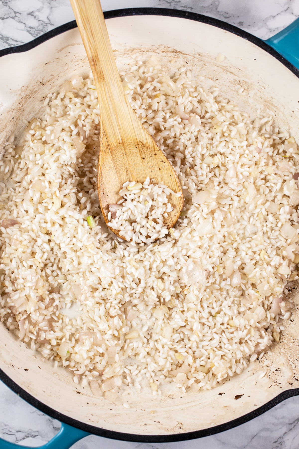 Garlic, shallots, and Arborio rice sautéed in skillet with wooden spoon.