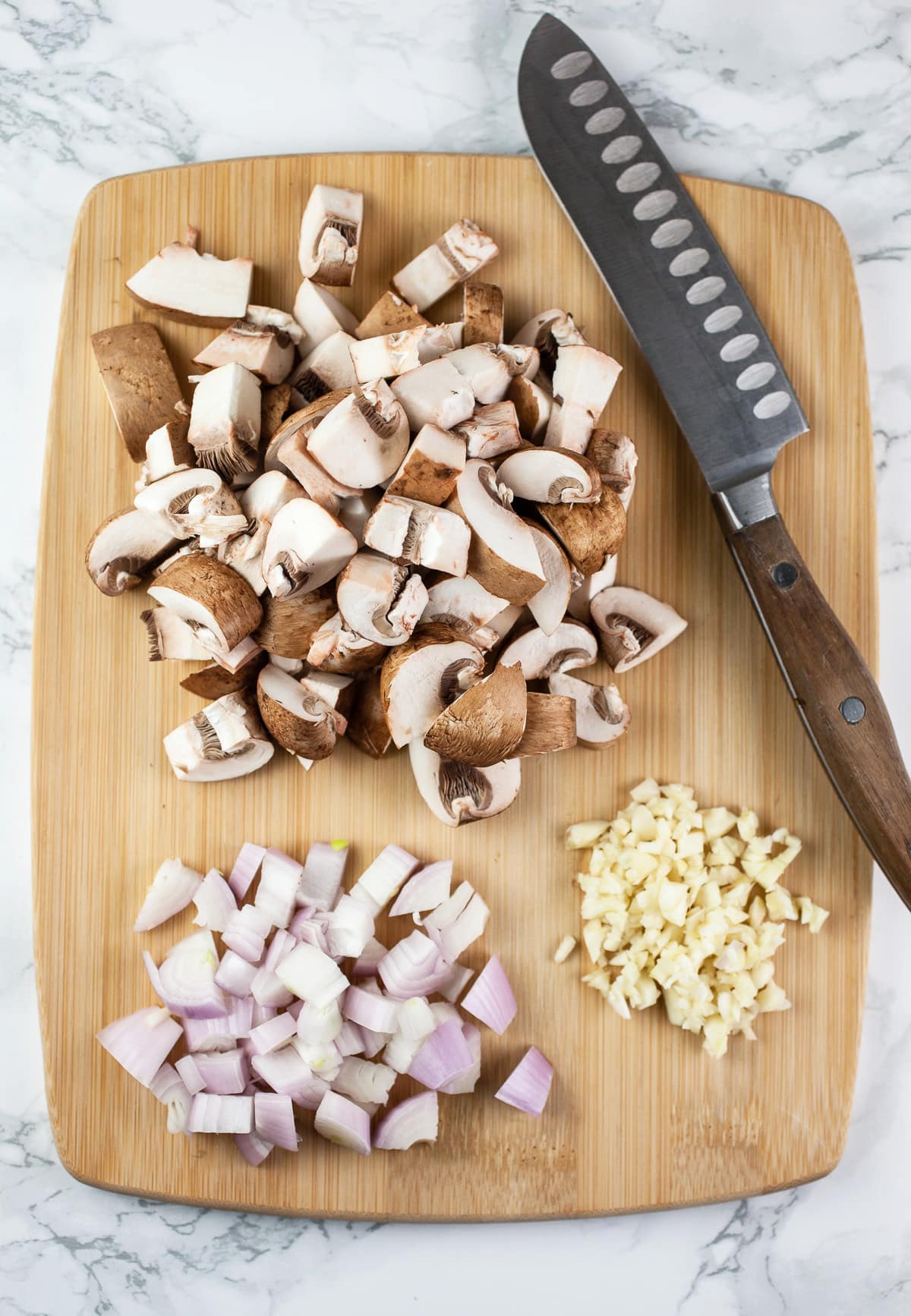 Minced garlic and shallots and diced mushrooms on wooden cutting board with knife.