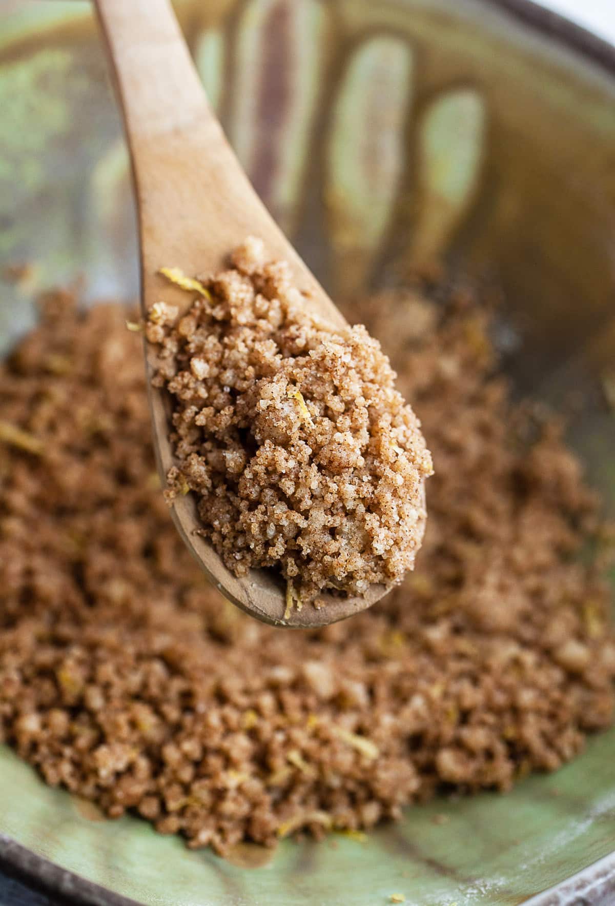 Spoonful of cinnamon streusel lifted from ceramic bowl with wooden spoon.