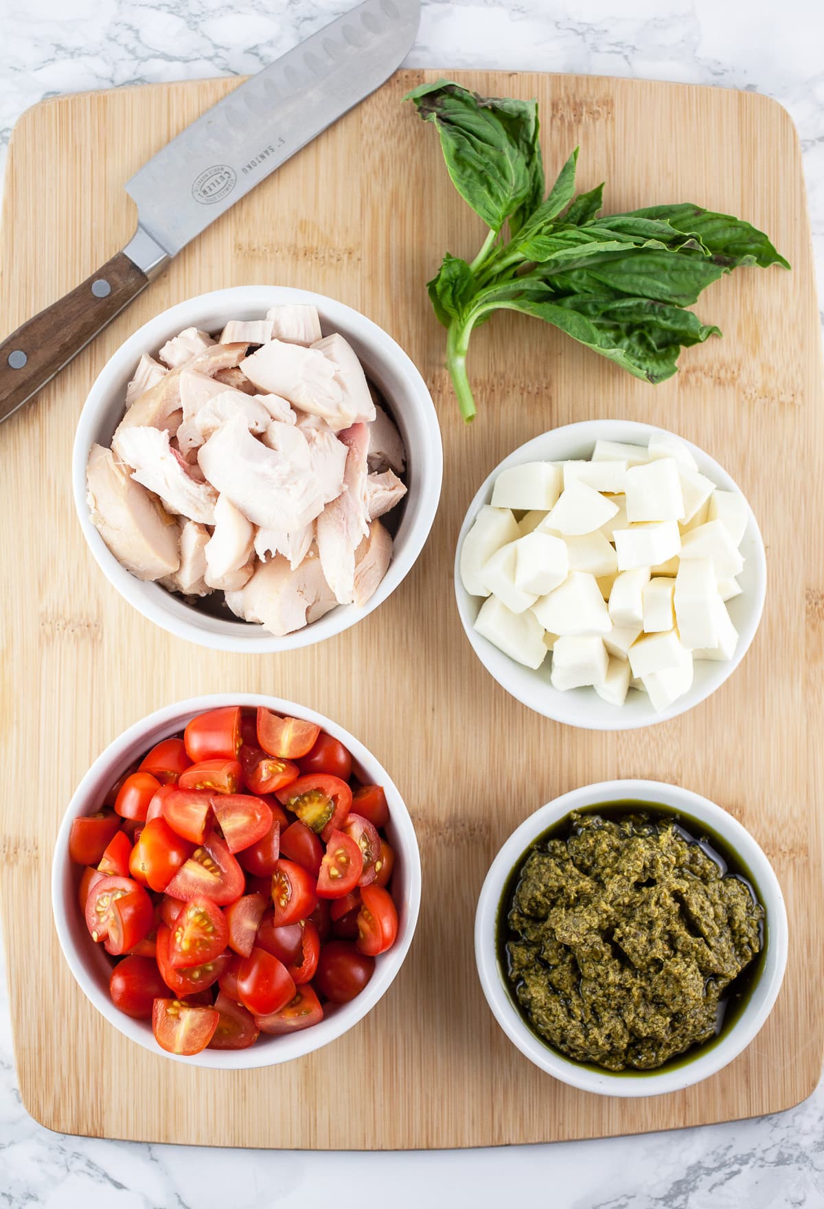 Chopped tomatoes, shredded chicken, mozzarella cheese, pesto, and fresh basil on wooden cutting board with knife.