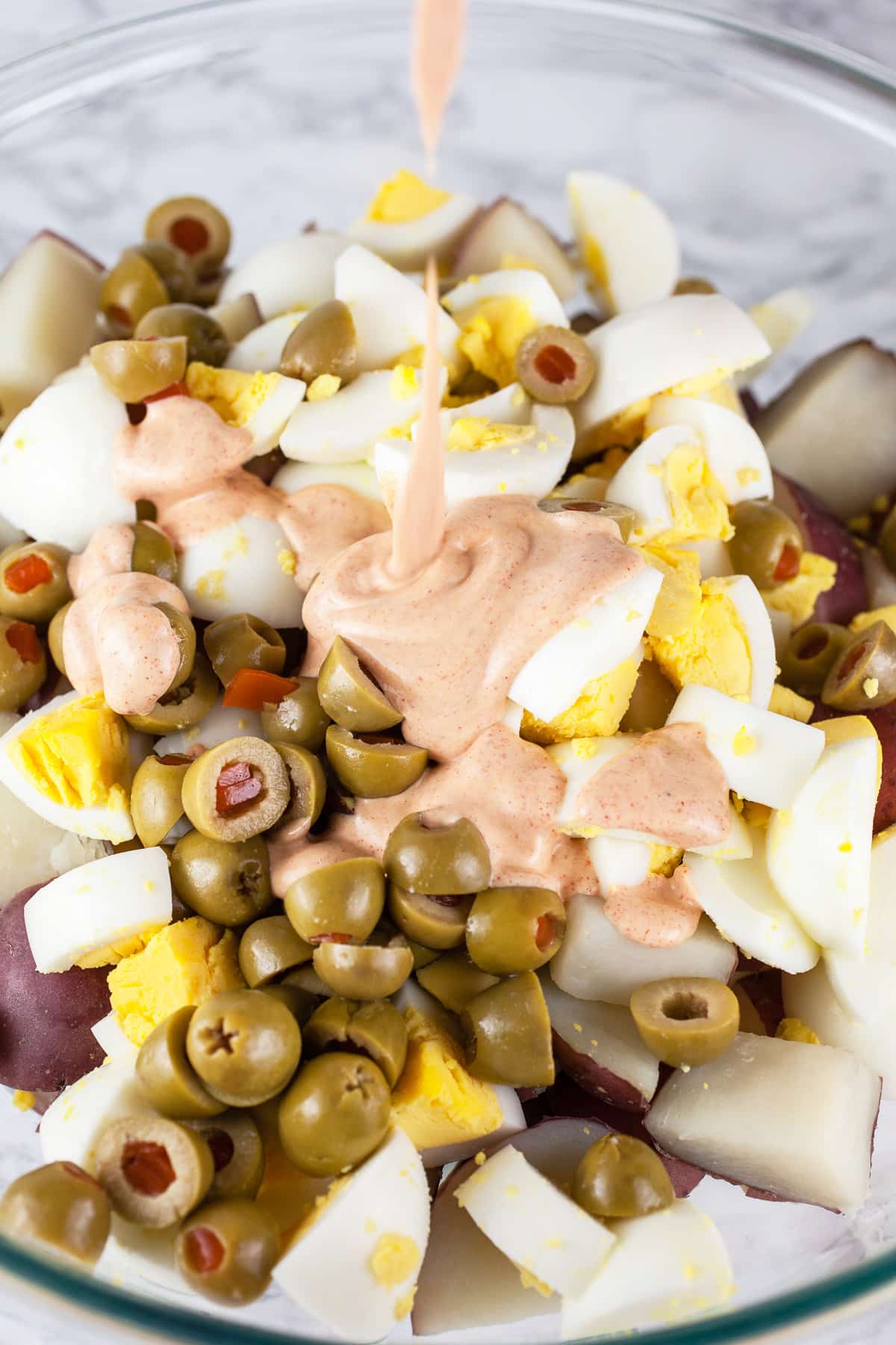 Dressing poured over potatoes, hard boiled eggs, and green olives in large glass bowl.