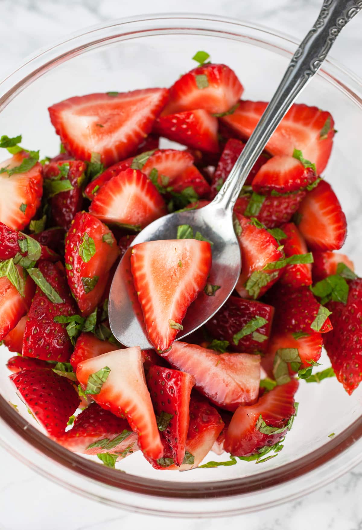 Sliced fresh strawberries tossed with lemon juice and mint in glass bowl.