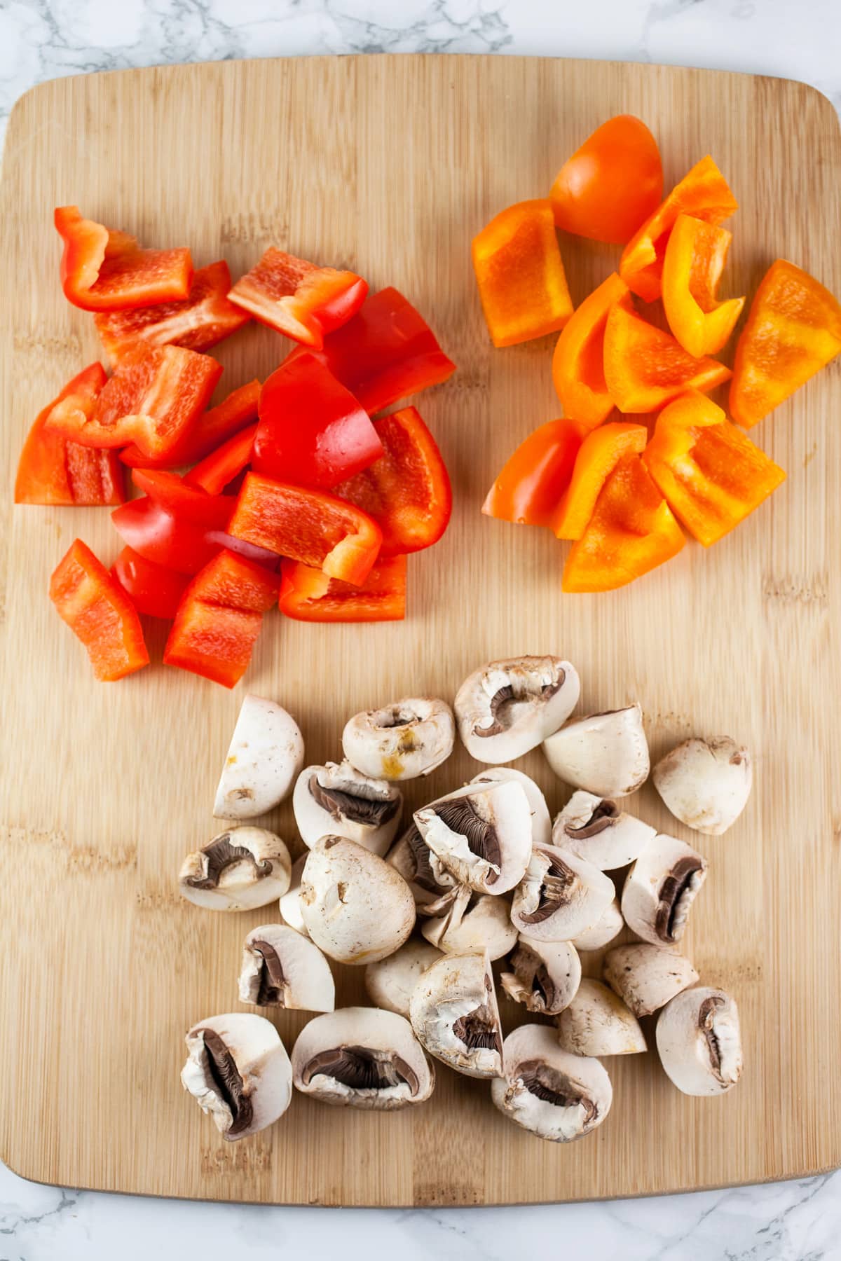 Chopped mushrooms and red and orange bell peppers on wooden cutting board.