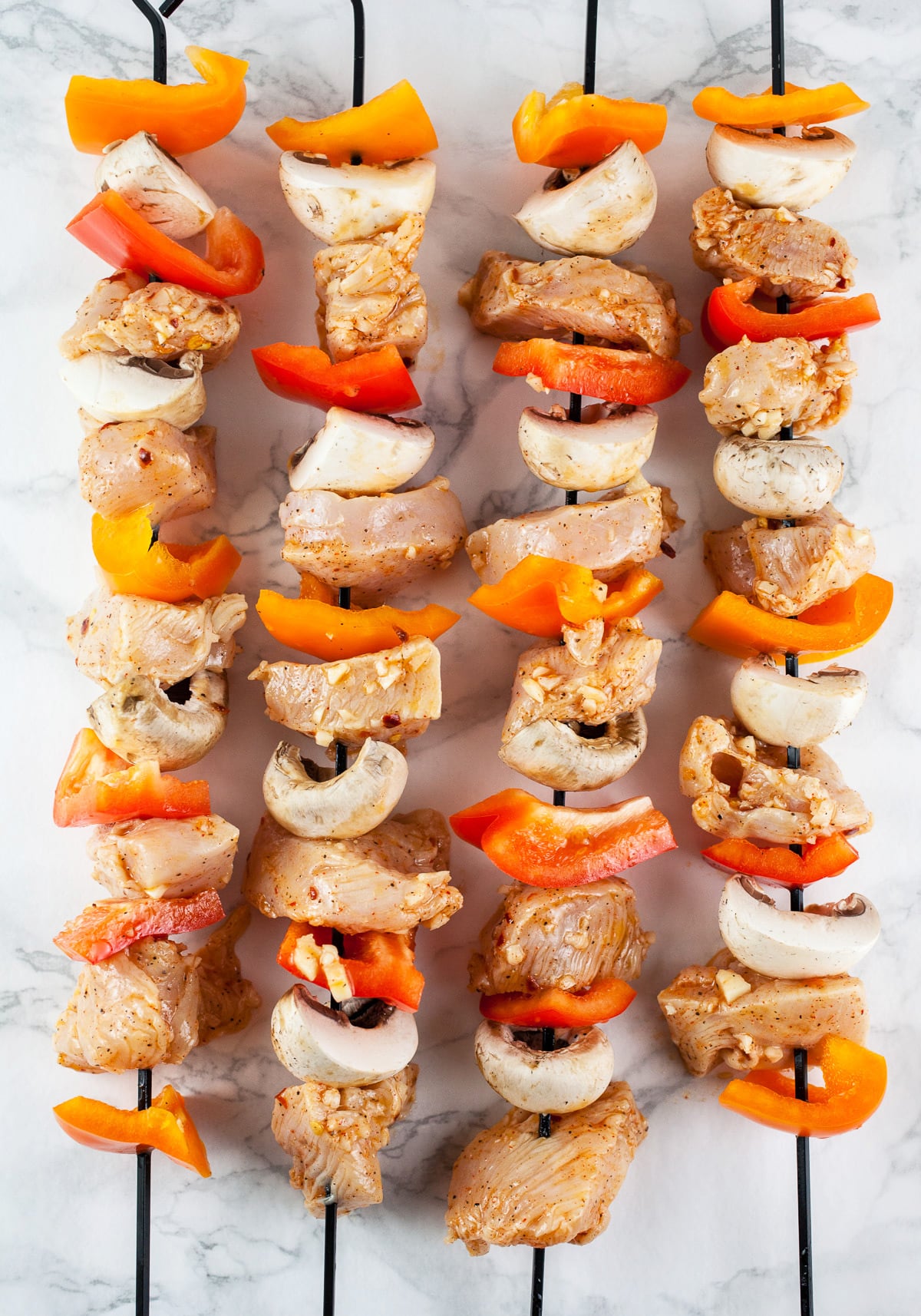 Raw chicken and uncooked bell peppers and mushrooms on metal skewers.
