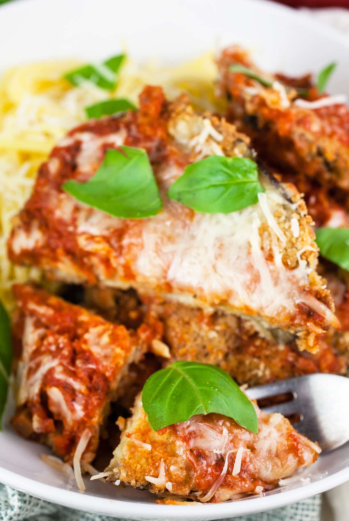 Eggplant parmesan cut with fork in white bowl with spaghetti noodles and basil.