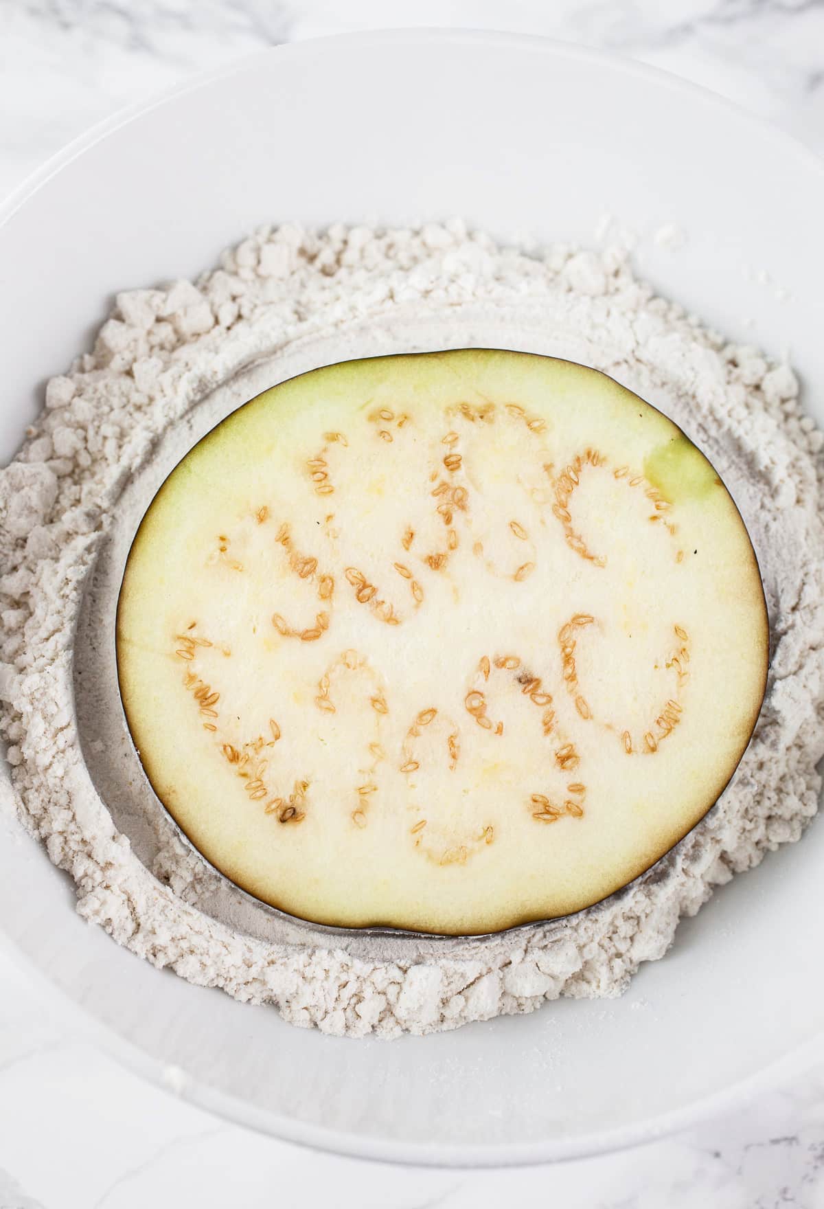 Eggplant slice in white bowl full of gluten free flour.