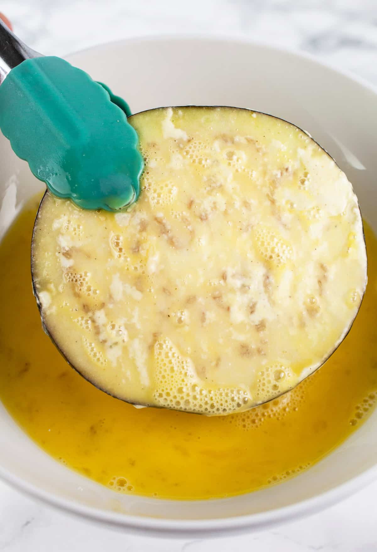 Eggplant slice dipped in egg mixture lifted from white bowl with spatula.