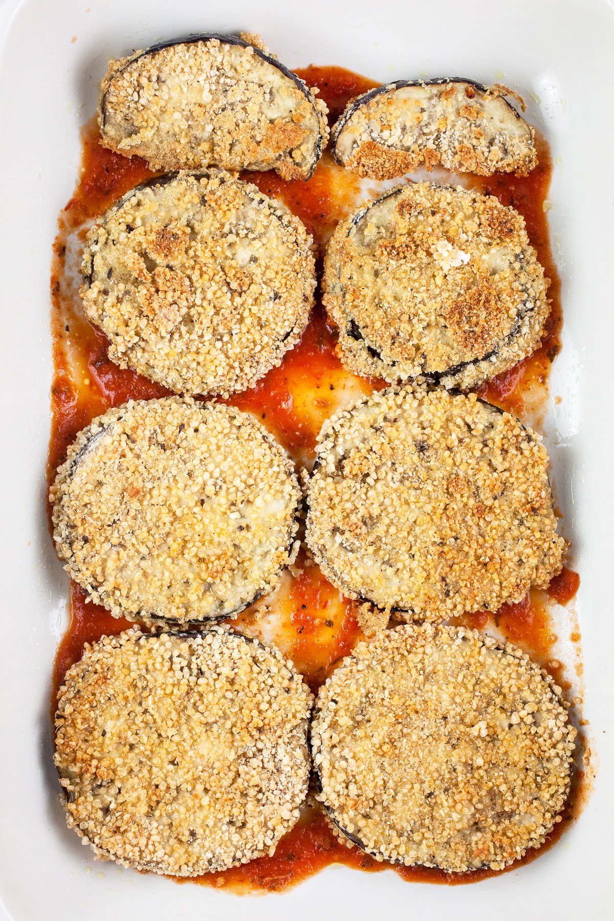 Baked eggplant slices and marinara sauce in ceramic baking dish.