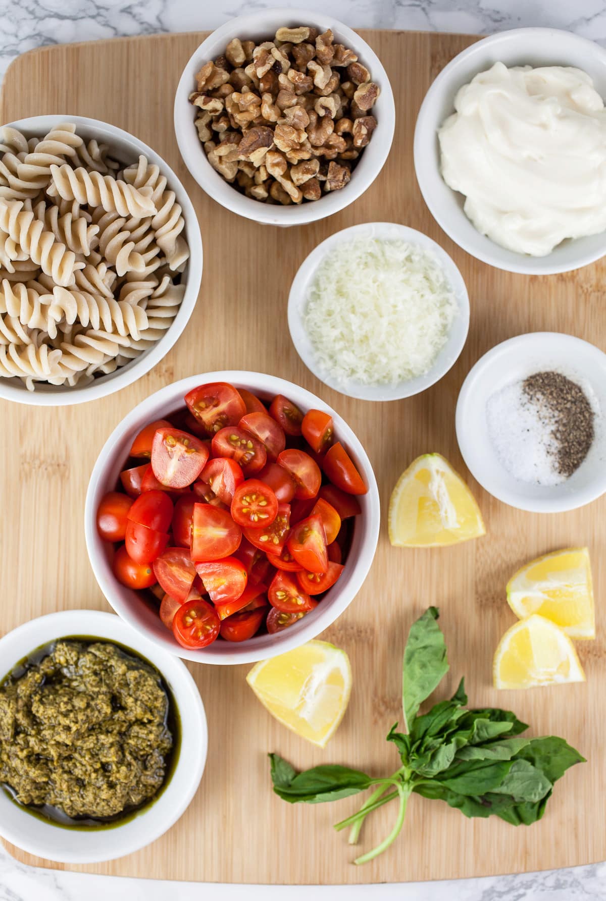 Jarred basil pesto, grape tomatoes, rotini pasta, walnuts, mayo, Parmesan, lemons, fresh basil, and salt and pepper on wooden cutting board.