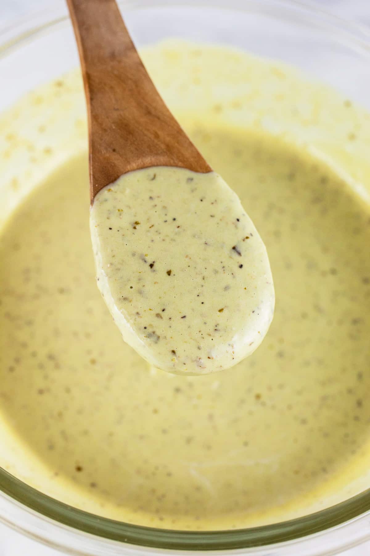 Spoonful of creamy pesto dressing lifted from small glass bowl on wooden spoon.
