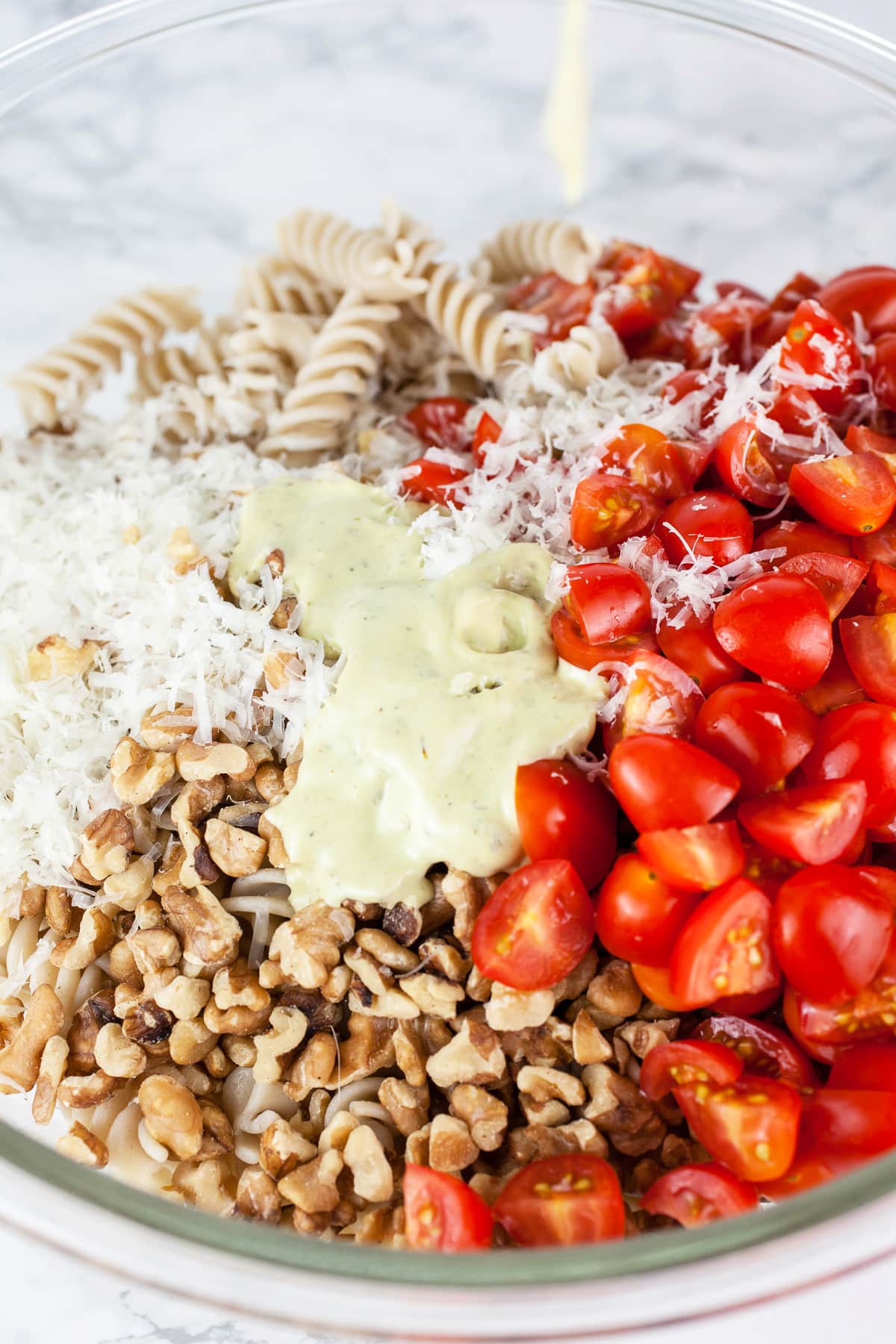 Creamy pesto dressing poured over pasta, tomatoes, walnuts, and Parmesan cheese in large glass bowl.