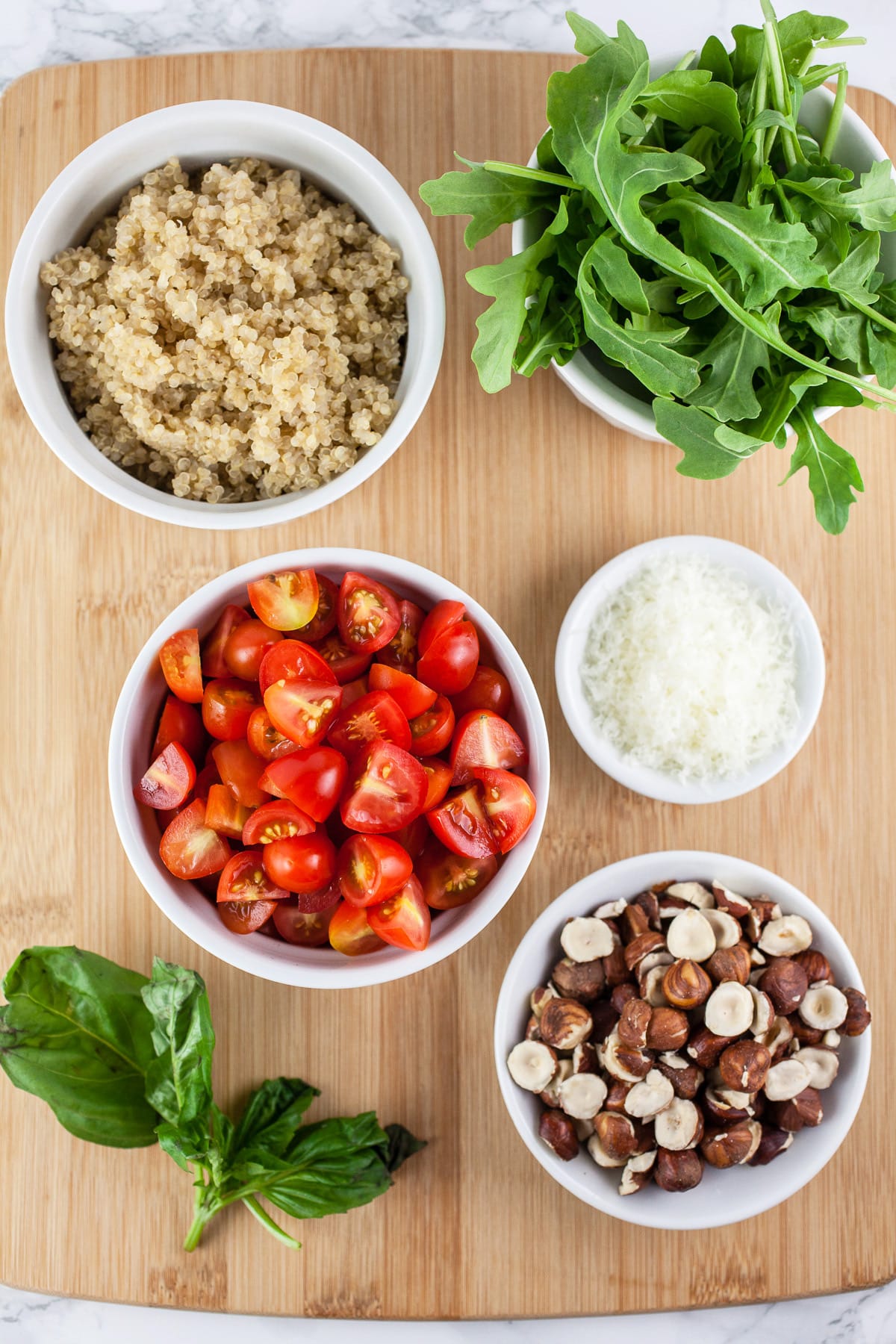 Basil, tomatoes, cooked quinoa, arugula, Parmesan cheese, and hazelnuts on wooden cutting board.