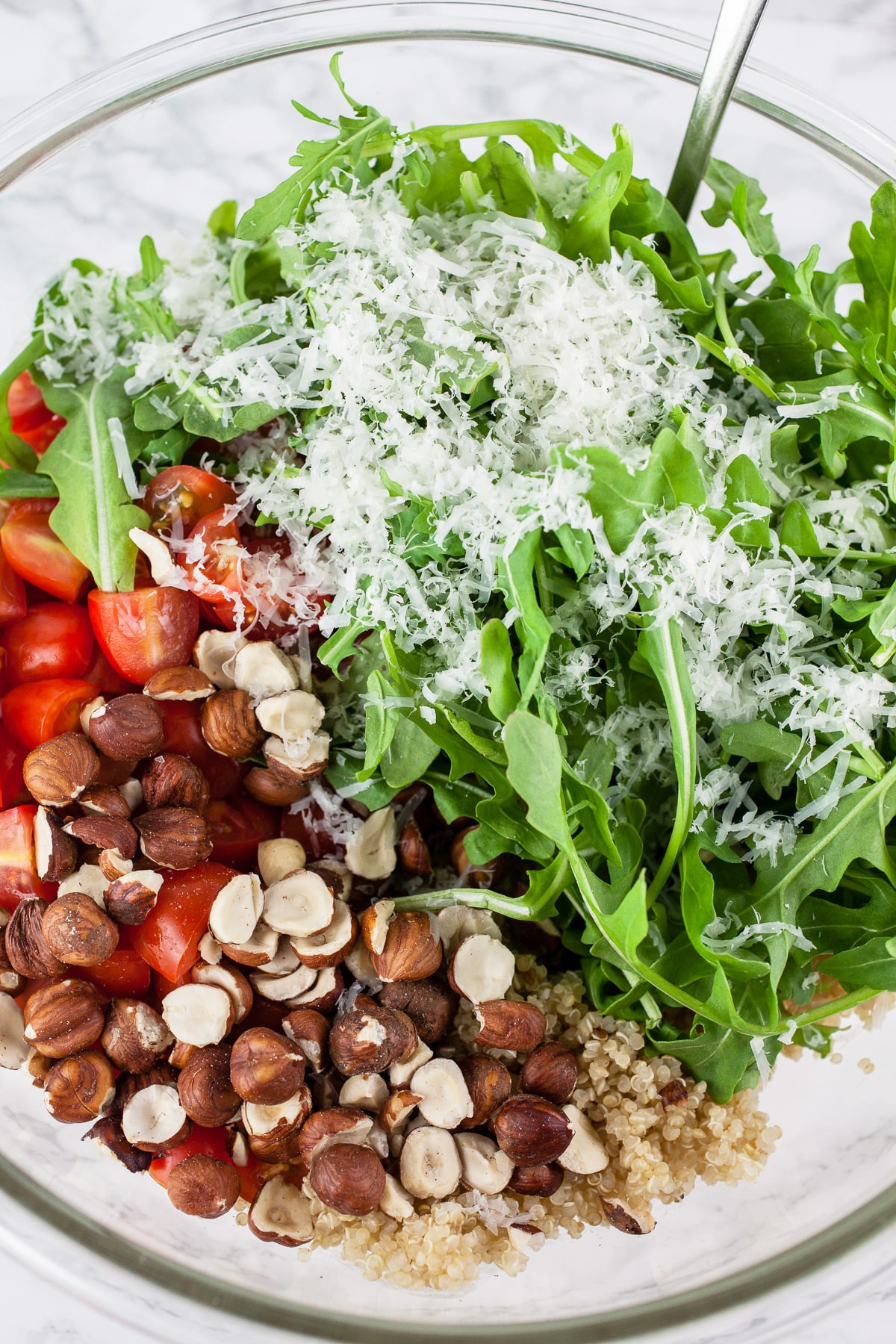 Quinoa, hazelnuts, tomatoes, arugula, and Parmesan cheese in large glass bowl with spoon.