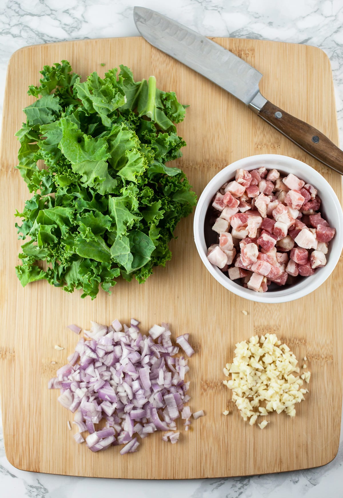 Minced garlic, shallots, chopped kale, and pancetta on wooden cutting board with knife.