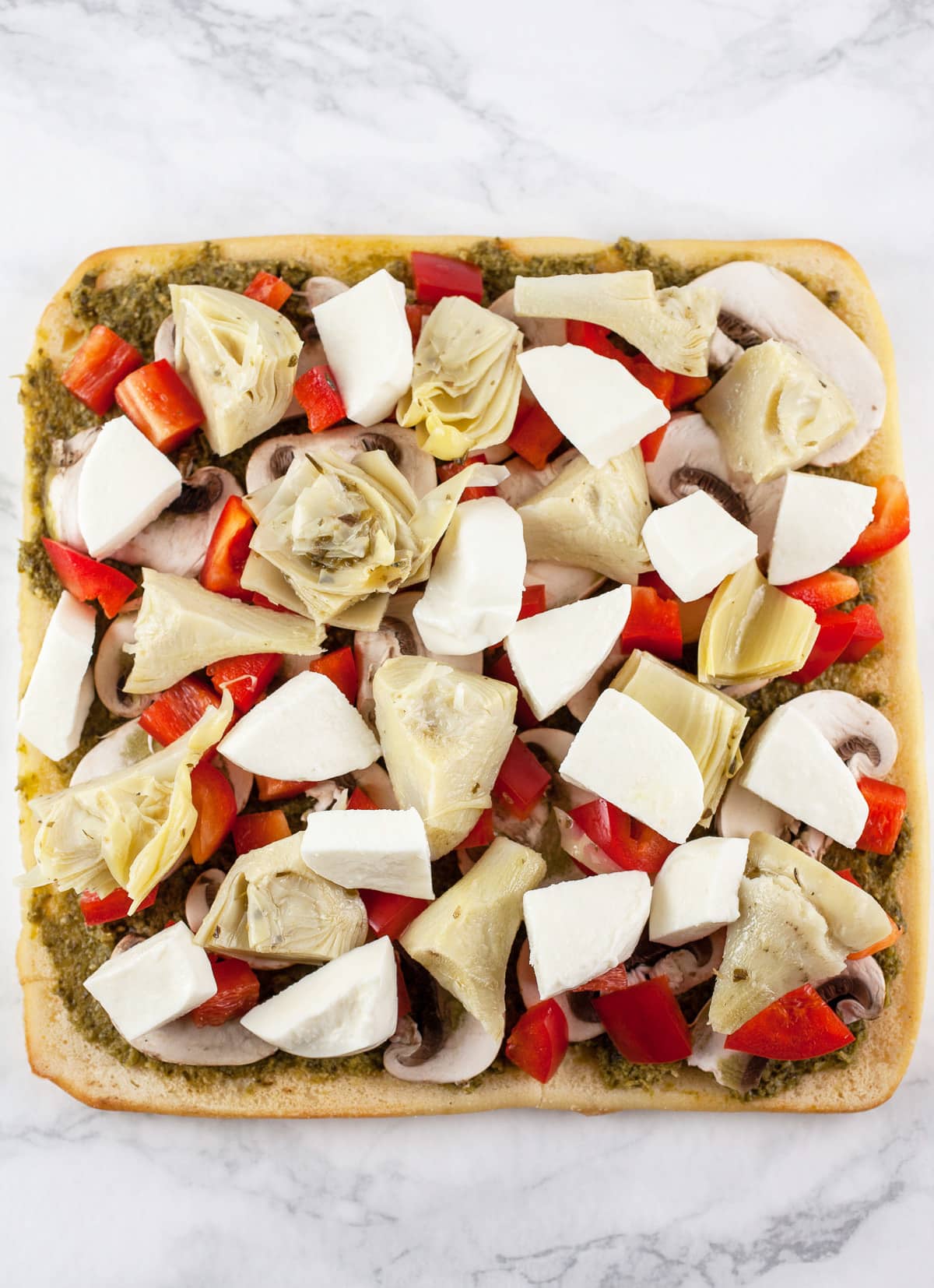 Pesto, red bell peppers, mushrooms, artichokes, and mozzarella cheese on uncooked flatbread.
