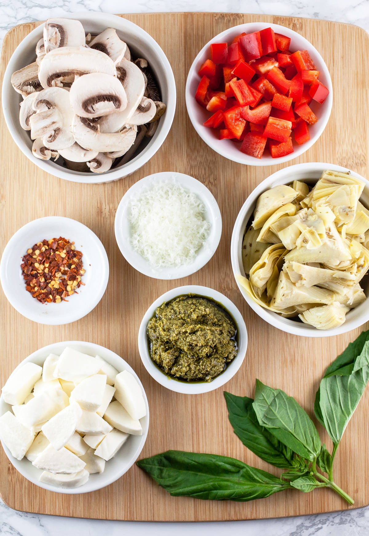 Mozzarella cheese, mushrooms, red bell peppers, marinated artichokes, basil pesto, red chili flakes, Parmesan cheese, and fresh basil on wooden cutting board.