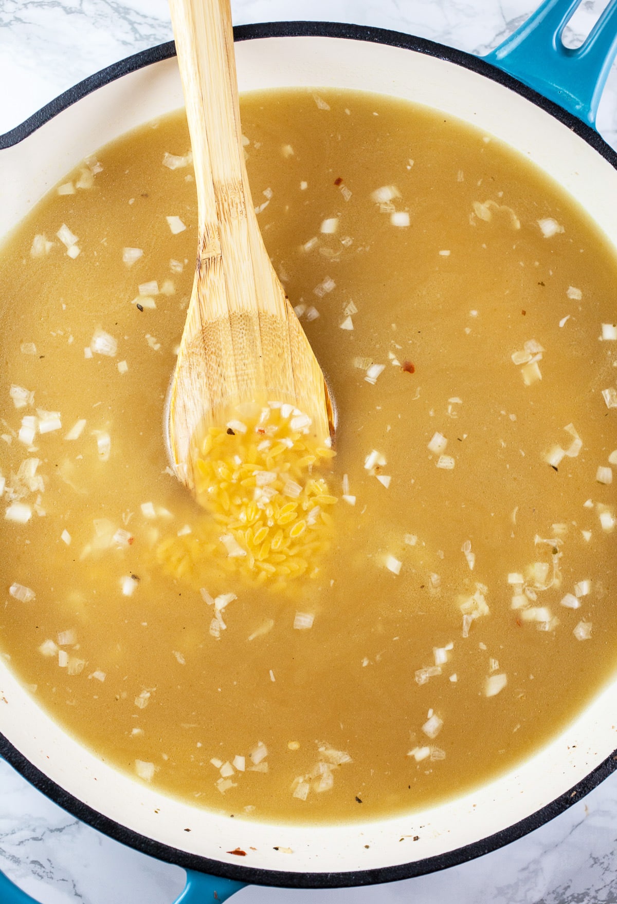 Garlic, shallots, orzo, and chicken broth in skillet with wooden spoon.