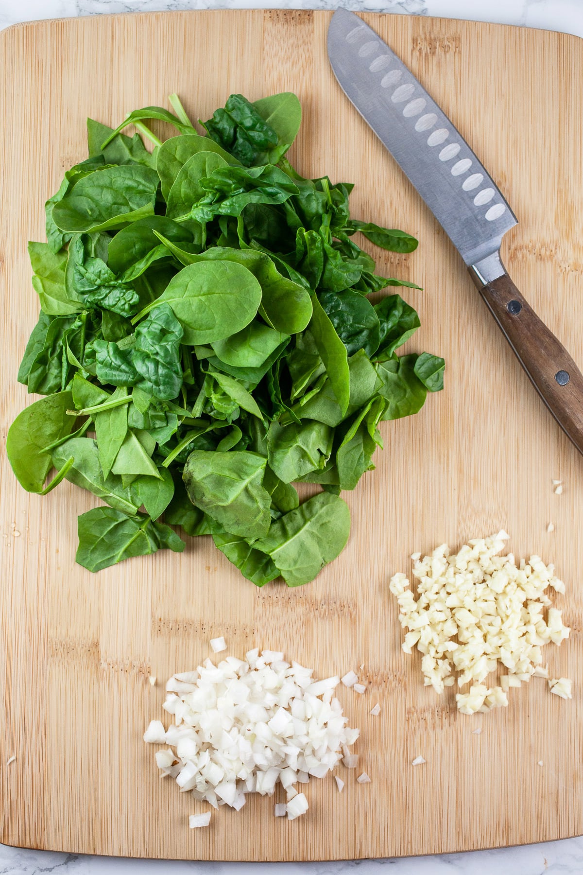 Minced garlic, shallots, and spinach on wooden cutting board with knife.