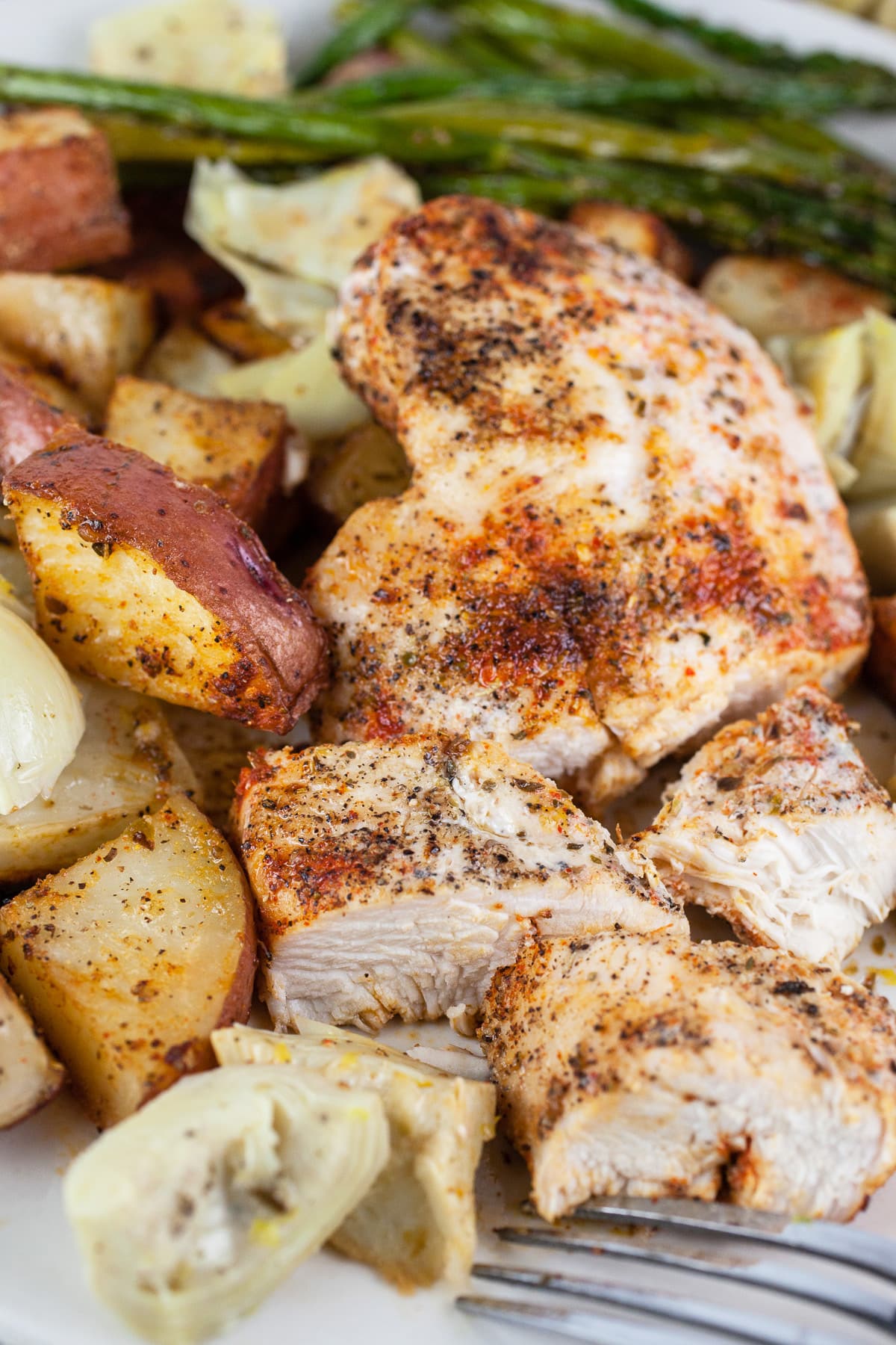 Mediterranean chicken and potatoes with roasted asparagus and artichokes on white plate with fork.