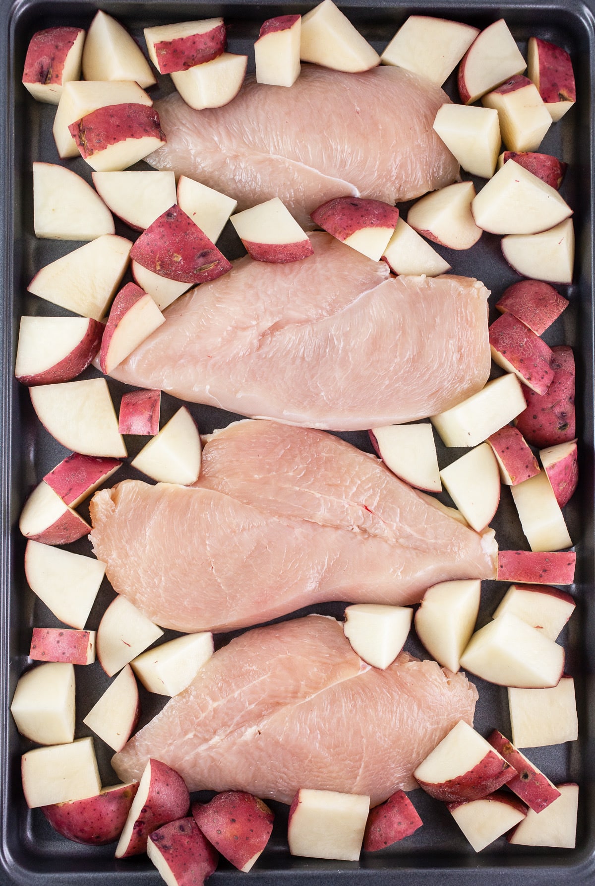 Uncooked chicken breasts and diced red potatoes on baking sheet.