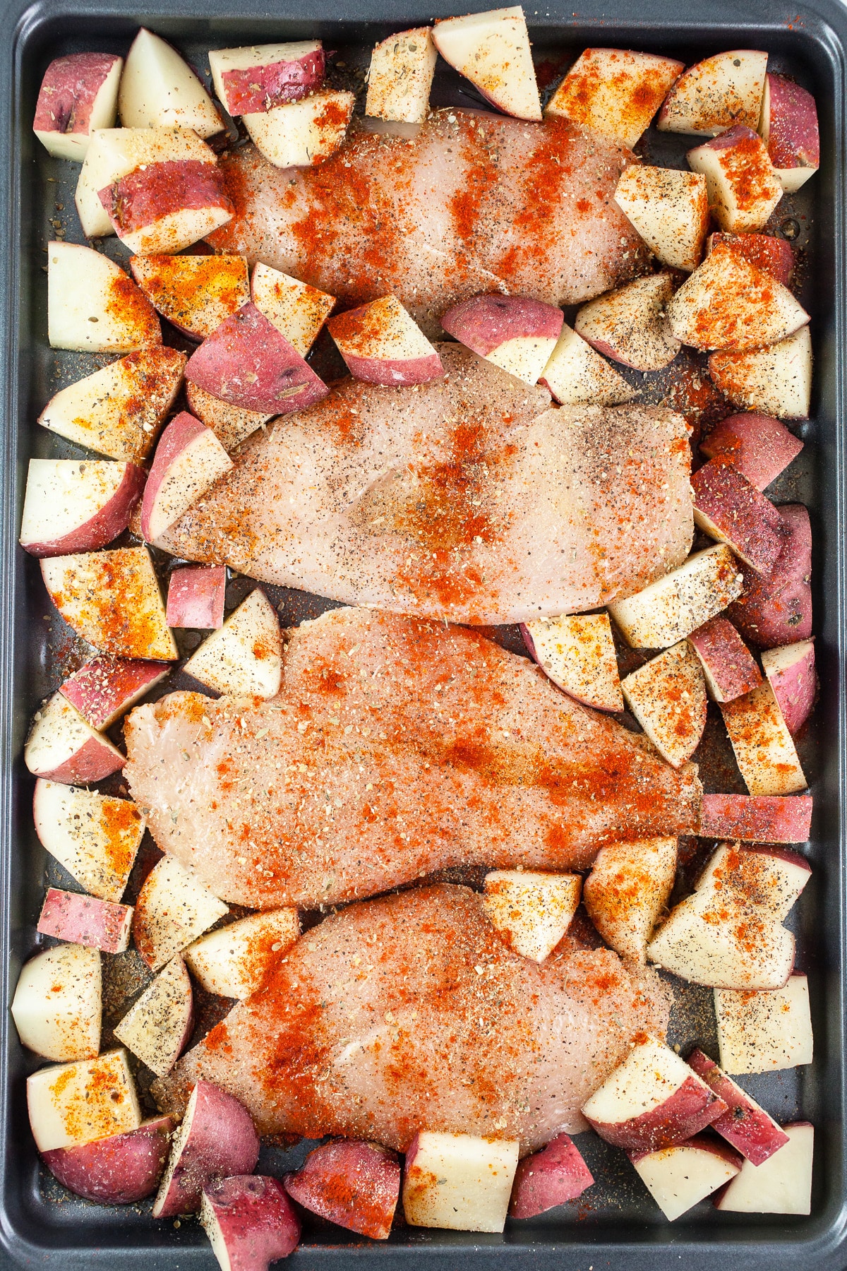 Chicken breasts and potatoes with spices on baking sheet.